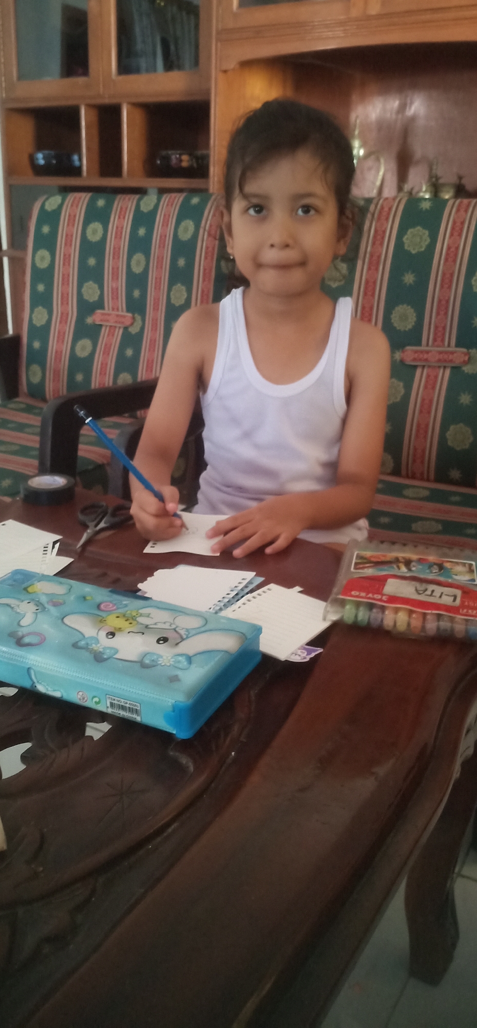 A young girl with dark hair and brown eyes is sitting at a wooden table, writing or drawing with a blue pencil. She is wearing a white tank top and is positioned in front of a sofa with a striped green and red pattern. On the table in front of her are several notebooks, a pencil case decorated with cartoon animals, a roll of black tape, and a pair of scissors. To her right, there is a package of colored pencils or markers. The background shows a wooden cabinet with shelves. The scene appears to be an indoor setting, likely a home. Based on the context of the city of Gending, Indonesia, this image likely depicts a domestic scene in a residential area within that city.