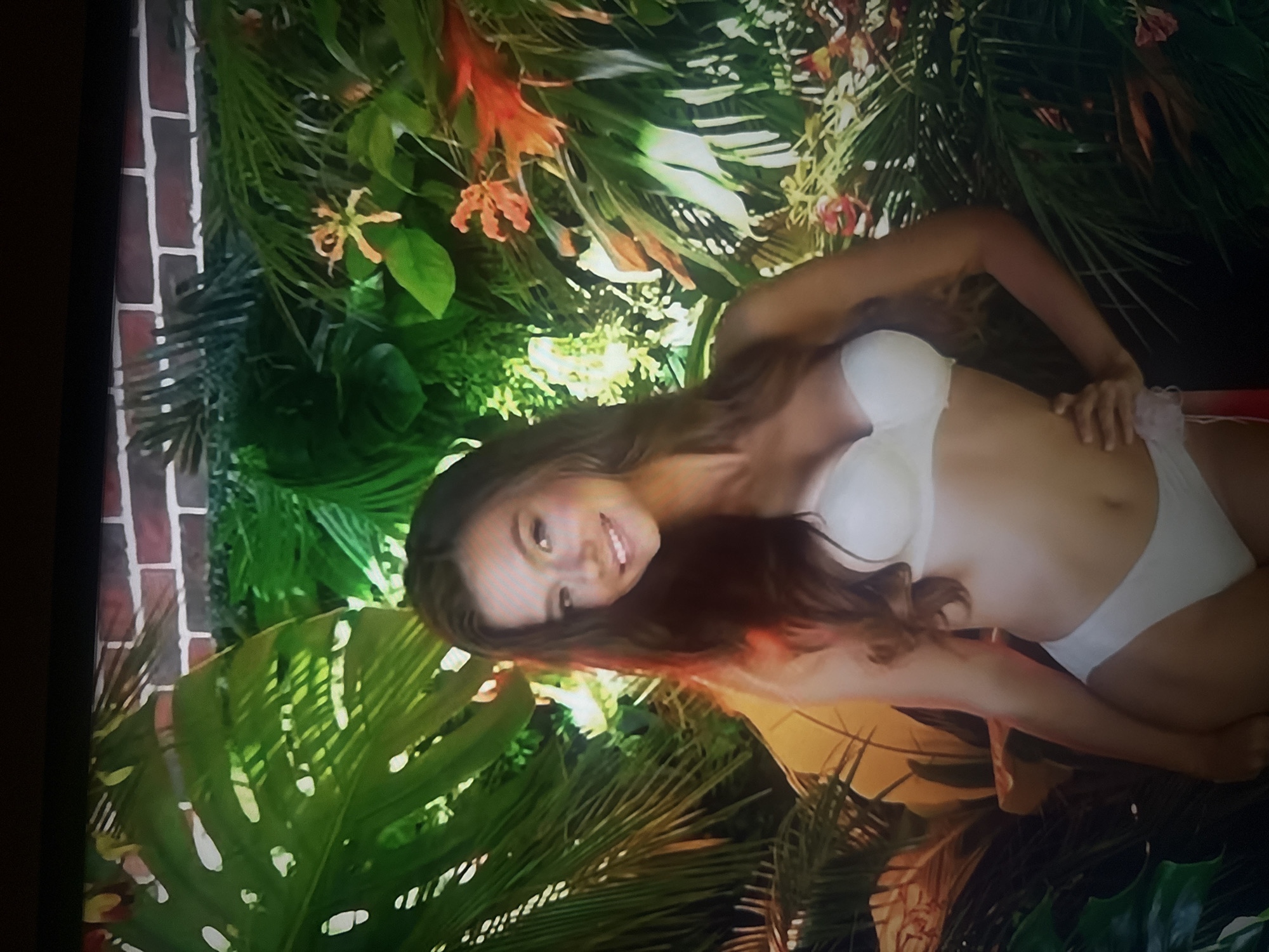 A young woman with long, wavy brown hair is pictured from the chest up, smiling at the camera. She is wearing a white strapless bra and white bikini bottoms. She is standing in front of a backdrop of lush green foliage, with some red and orange tropical flowers interspersed. The backdrop also features a brick wall pattern at the top. The lighting is bright, with a warm glow illuminating the woman. The overall impression is that of a tropical setting. The location is not identifiable as Herzliya, Israel.
