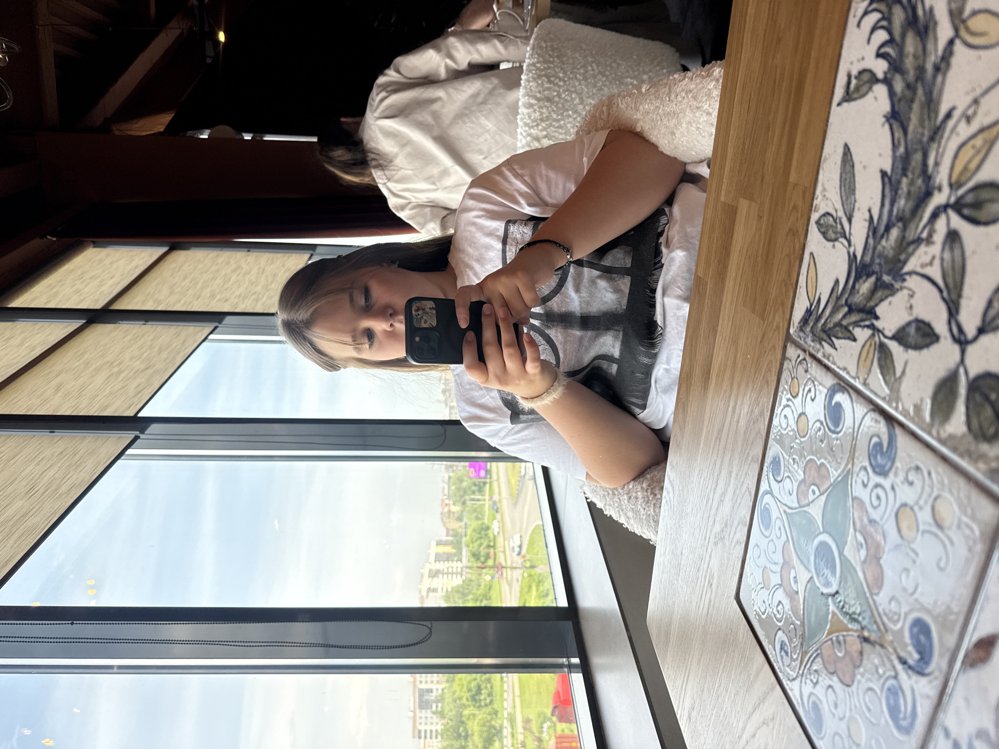 A young woman with light brown hair is seated by a large window, holding a black smartphone in her hands. She is looking at the screen of the phone. She is wearing a white t-shirt. The scene appears to be inside a restaurant or cafe, as evidenced by the patterned tile table and the presence of other people in the background. The large window provides a view of a city, which is Veliky Novgorod, Russia. The outside view shows buildings, trees, and a road with car traffic. The weather appears to be bright and overcast.