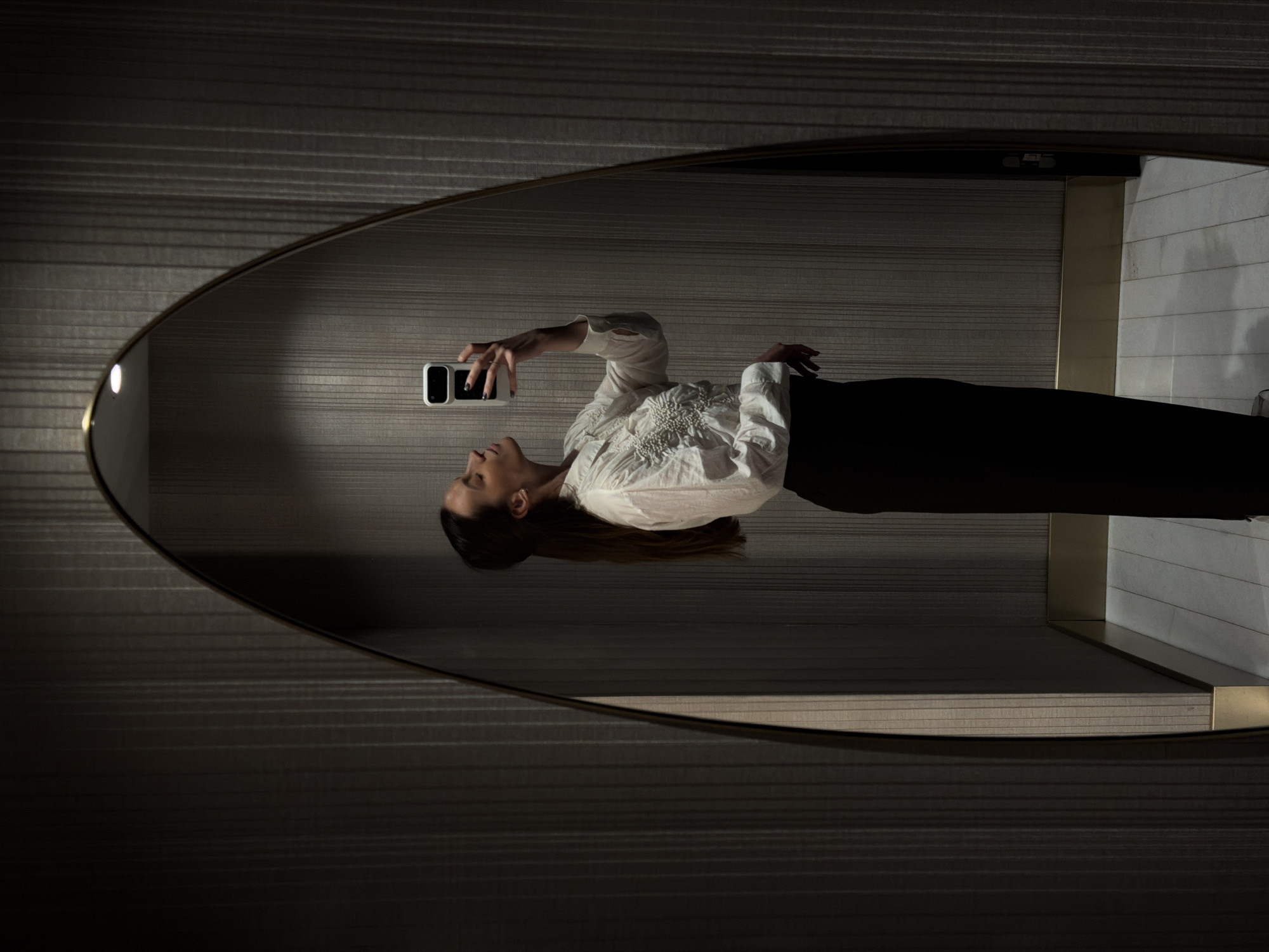 A woman is taking a selfie in an oval mirror. The mirror is positioned against a wall covered with horizontal lines. The woman is wearing a white blouse with embellishments and black pants. Her hair is long and brown. Her left hand is holding a smartphone in front of her face. The mirror's frame is bronze. The location is Budva, Montenegro.