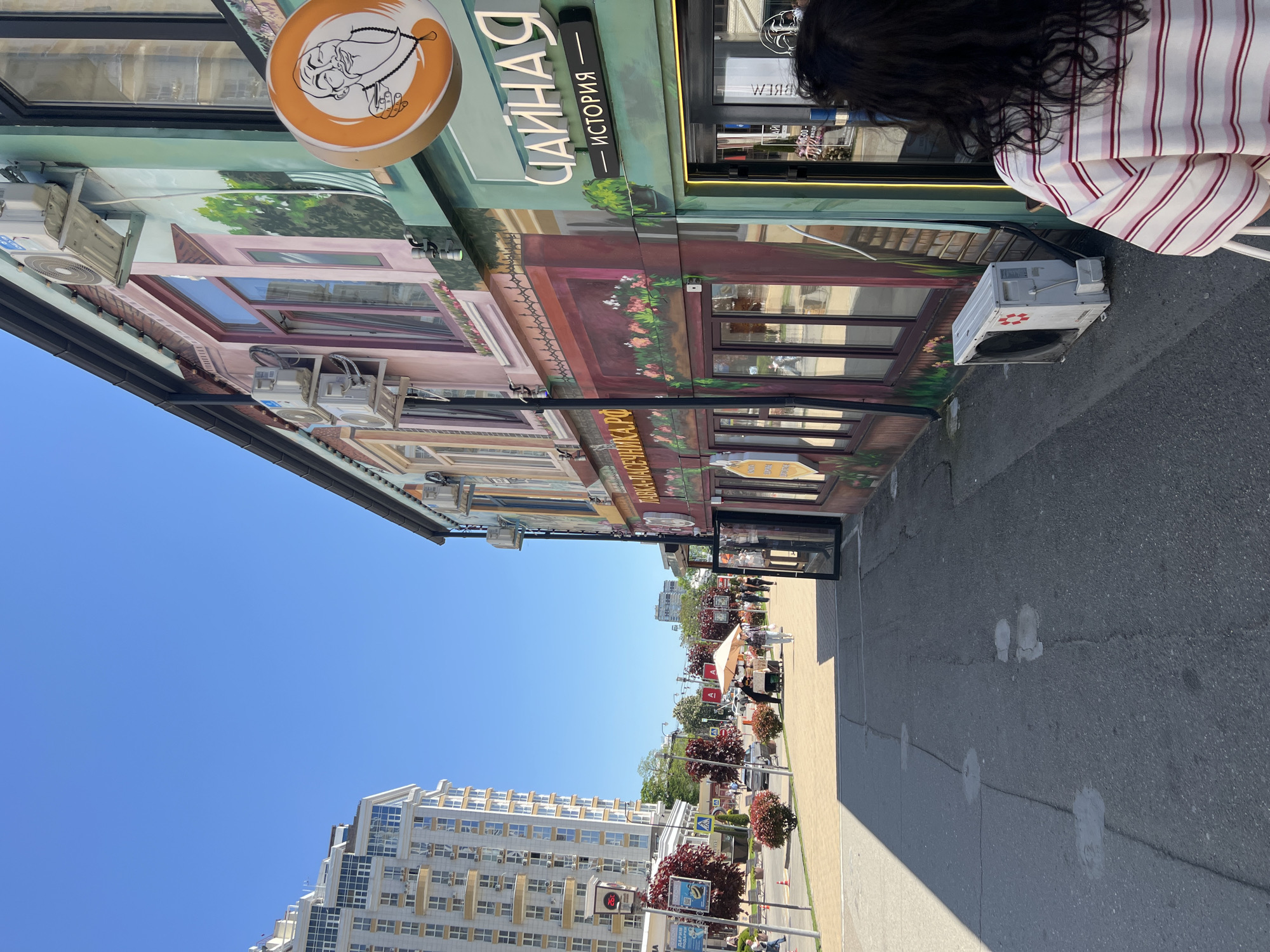 The image captures a street scene in Krasnodar, Russia. A building with a colorful mural depicting building facades stretches across the upper left and top of the frame. Signs in Russian identify a tea shop ("ЧАЙНАЯ ИСТОРИЯ") with a logo of a man with a tea set. Numerous air conditioning units are attached to the building. A sidewalk runs along the right side of the frame, leading towards the center. People are visible walking along the sidewalk. A modern apartment building stands across the street. The sky is clear and blue.
