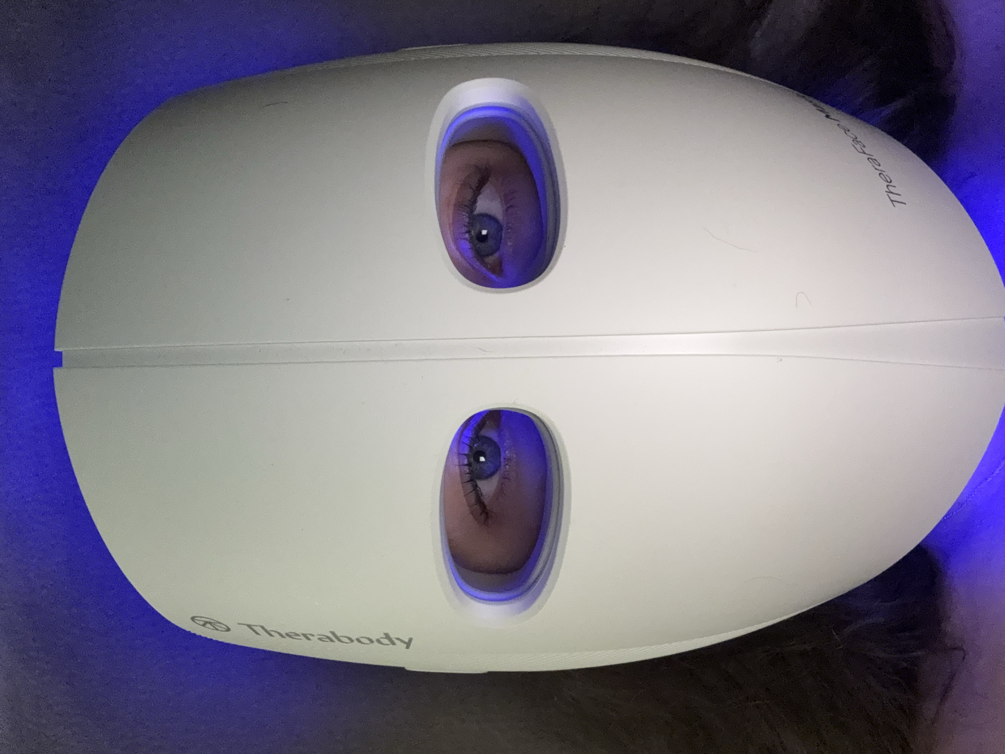 The image shows a person wearing a Therabody TheraFace Mask. The mask is white with two eye holes through which the person's eyes are visible. A blue light illuminates the mask. The surroundings are blurry, but suggest dark hair and a soft surface. There is no indication of the location being Tel Aviv, Israel or any other specific location.
