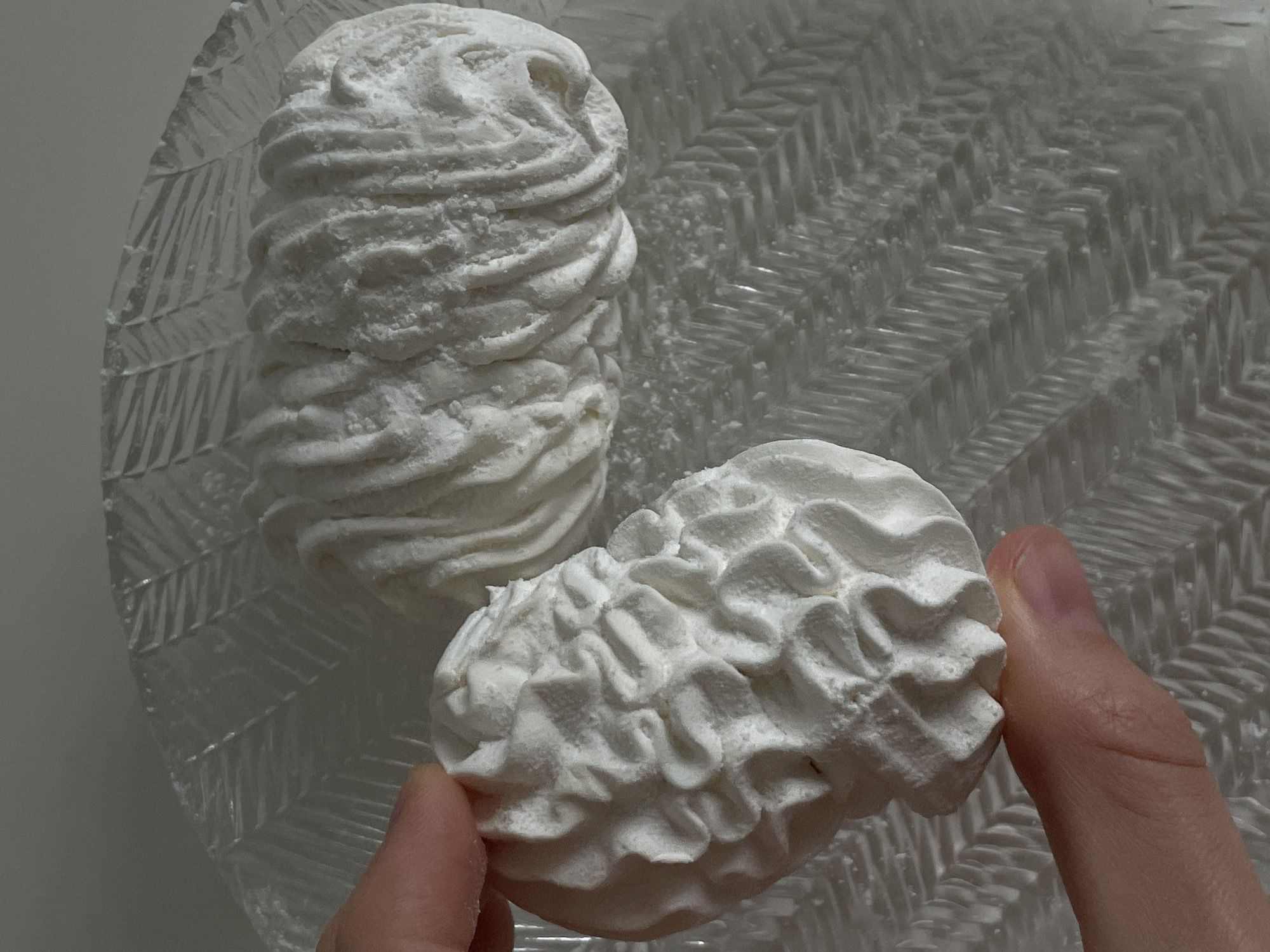 A person's thumb and forefinger hold a white, fluffy, meringue-like confection with intricate, swirled ridges. The confection is placed on a clear glass plate with a raised, geometric pattern. Another similar confection rests on the plate behind the one being held. The setting appears to be indoors, with neutral-colored walls in the background. There is no discernible information about the location in Nidda, Germany.