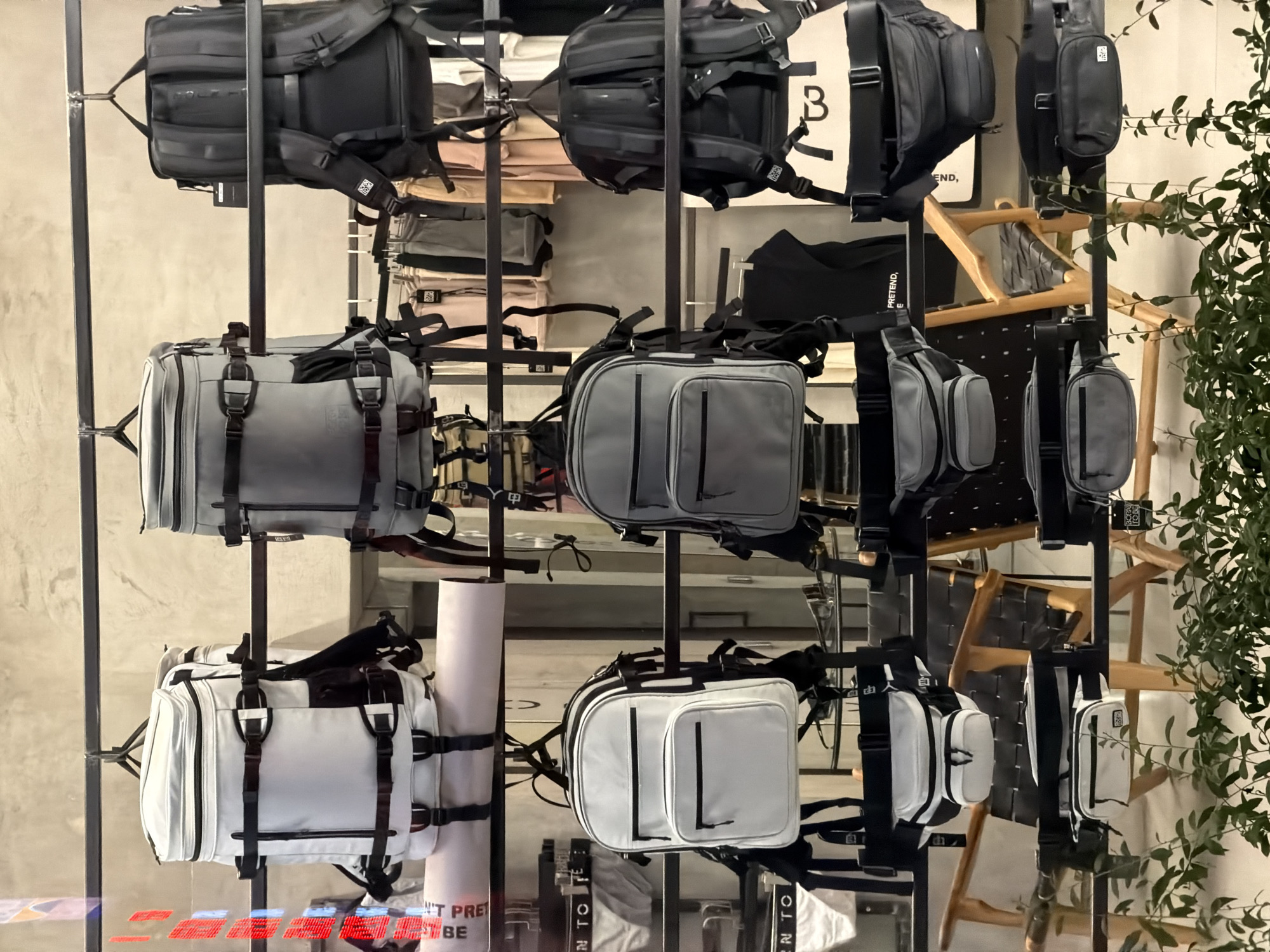 The image shows a display of bags in a shop. The bags are organized on a black metal grid, with varying styles and colors, mostly black, grey, and white. The bags include backpacks, duffel bags, and smaller crossbody bags. A sign with letters and symbols is visible in the foreground. There is a roller on the rack that reads "DON'T PRETEND TO BE". The background shows a concrete wall and wooden furniture. A green plant is seen in the right side of the frame. The sign "BORN TO PRETEND" is visible in the background. A small tag is visible on each bag. The bags are displayed on a rack. The location is North Kuta, Indonesia.
