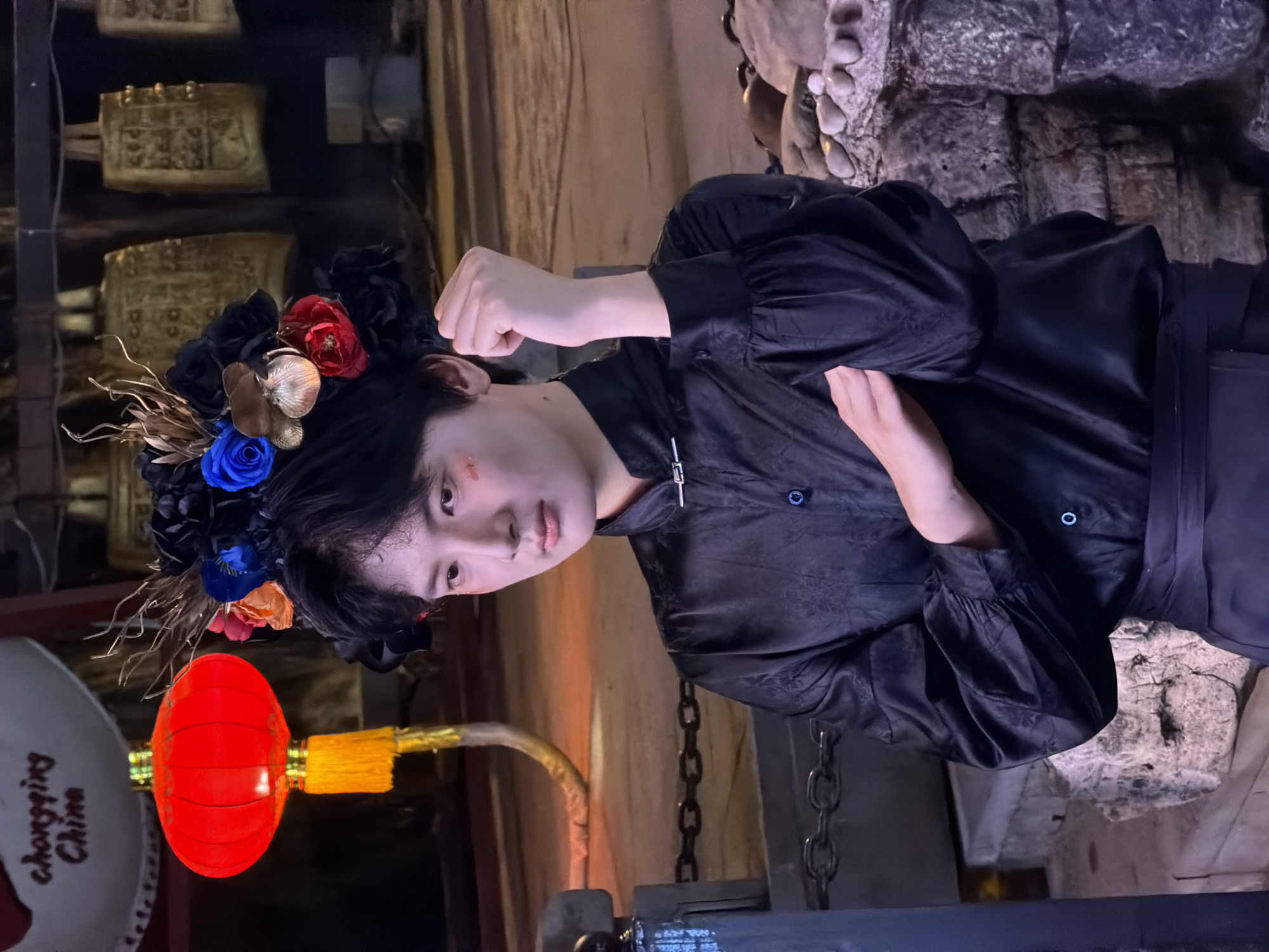 This media file features a young person with dark hair adorned with a floral headdress. The headdress includes black, red, blue, and orange flowers, along with gold metallic accents. The person is wearing a black silk shirt with intricate embossed patterns and puffed sleeves. They are positioned with their body angled and their left arm raised, fist clenched, in a striking pose. A notable detail on their face is a small reddish mark on their cheek.
In the background, there are traditional Chinese bronze bells, some of which are partially obscured. A red, oval-shaped lantern hangs to the left of the person, with visible text on a white circular object behind it that appears to say "Chongqing China". The setting suggests an indoor or covered outdoor space with a rustic or historical aesthetic, featuring stone-like textures and metal chains. The lighting is dim, indicating it might be evening or a dimly lit indoor environment. The overall mood conveyed by the pose and attire could be interpreted as dramatic or theatrical.