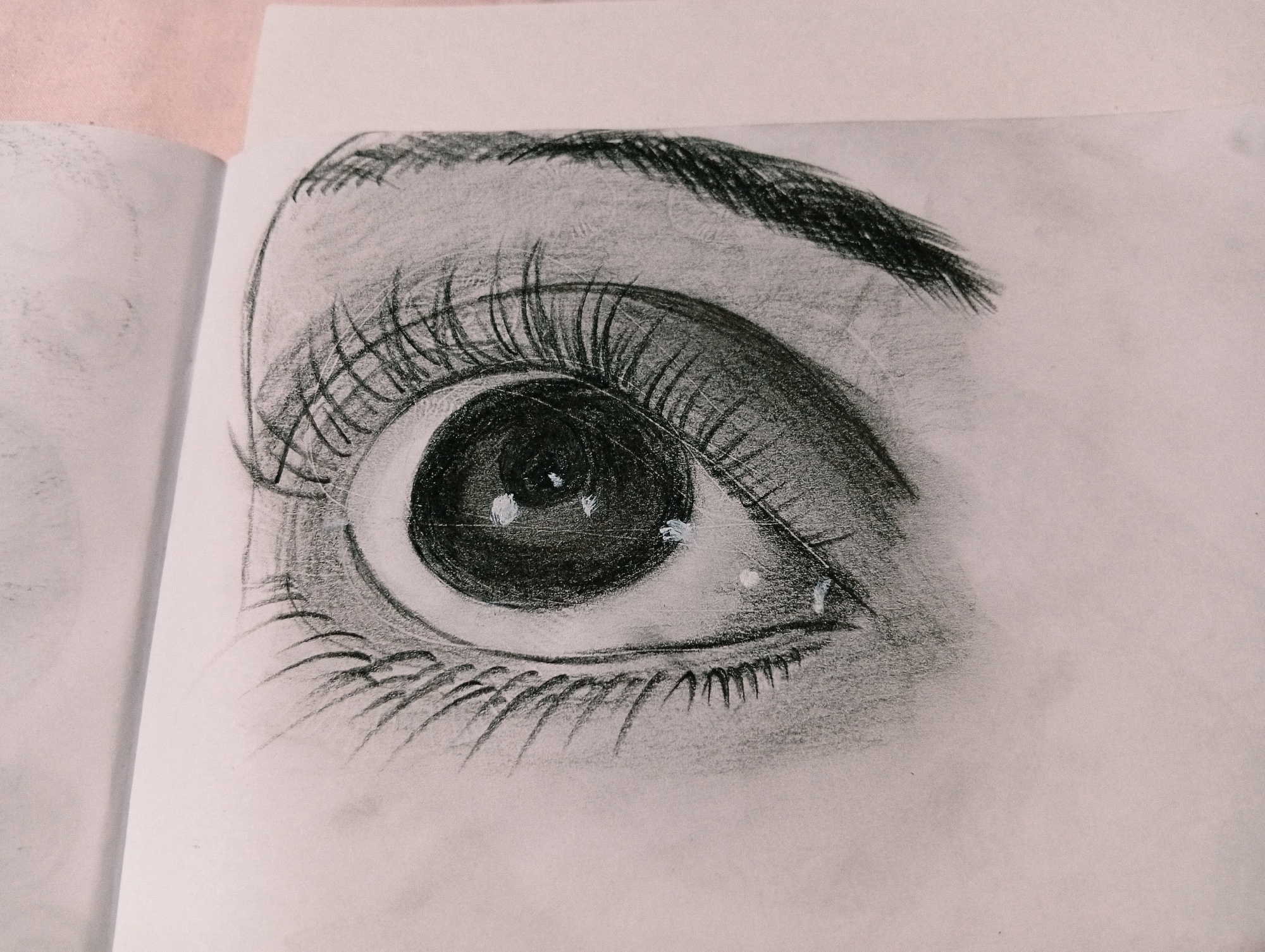 This is a charcoal drawing of a human eye, rendered on a sheet of paper. The drawing captures the detail of the eyelashes, iris, pupil, and eyebrow with varying shades of gray. There are highlights on the iris, suggesting light reflection. The paper appears to be from a sketchbook, as indicated by the presence of another drawing on the left page and the binding on the left side. The background is a light pinkish hue, possibly fabric, and the surface is slightly textured. There is no information about the location in San Antonio de Los Altos, Venezuela, or any people, objects, actions, or interactions in the scene.