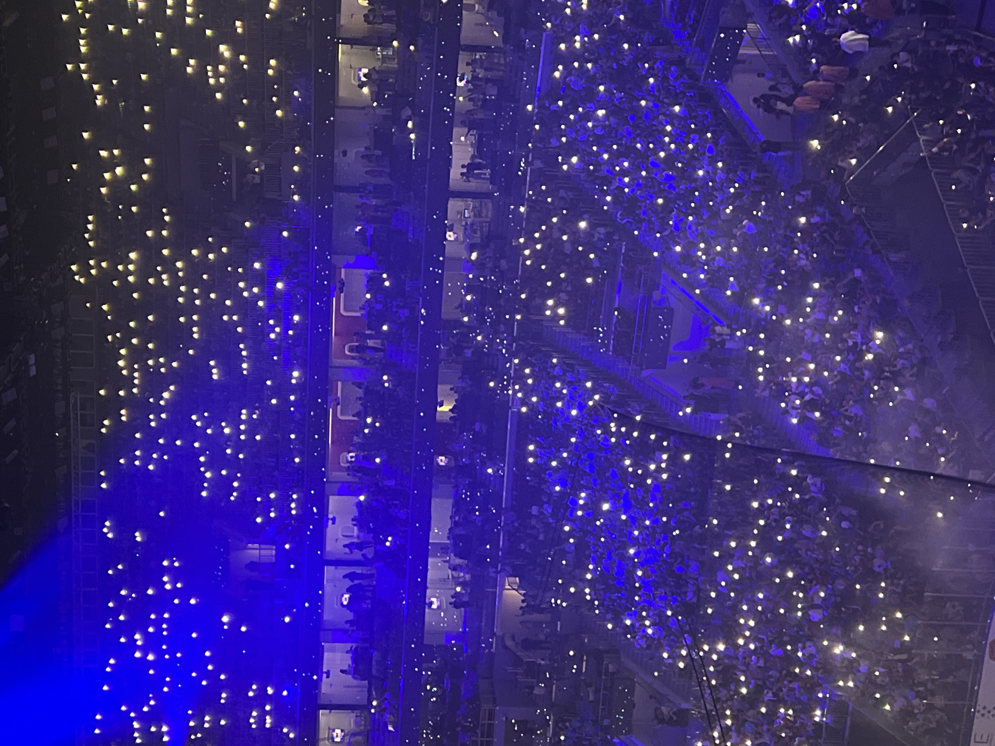 The image captures a wide, aerial view of a large indoor stadium filled with a crowd of people, illuminated by numerous handheld lights. The lighting is predominantly blue, with bright white pinpricks of light emanating from the audience's phones or other light sources, suggesting a concert or sporting event. Several tiers of seating are visible, occupied by dense gatherings of people. Sections of the stadium's upper levels also show people, some standing and gesturing. In some of the upper boxes, small groups of individuals are discernible. The overall atmosphere appears to be one of excitement and shared experience. The scene is set in a modern stadium environment, with visible architectural elements of the seating areas and structural supports. There is no indication of the city of Saint Petersburg, Russia, in the image itself; this information is external to the visual data.