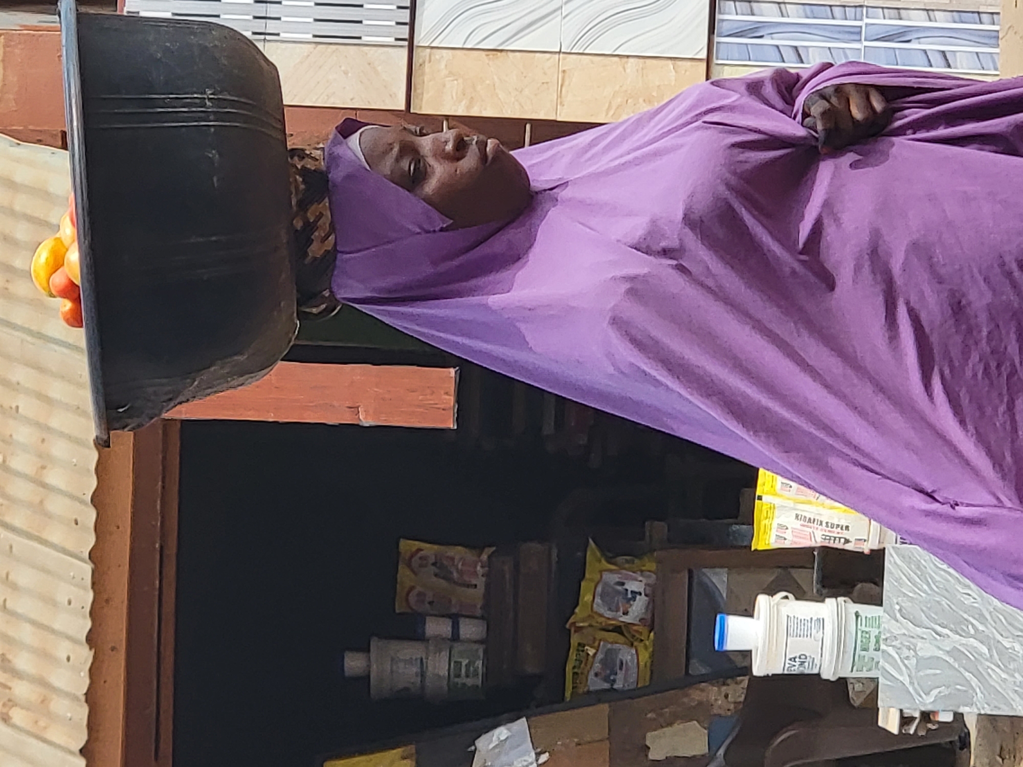 A woman wearing a purple garment and headscarf is lying on a surface in what appears to be a market setting. On her head is balanced a large, black bowl containing what appear to be tomatoes and oranges. Behind her is a cluttered stall containing boxes, tiles, and various small packages. The location is likely in Maiburiji, Nigeria.
