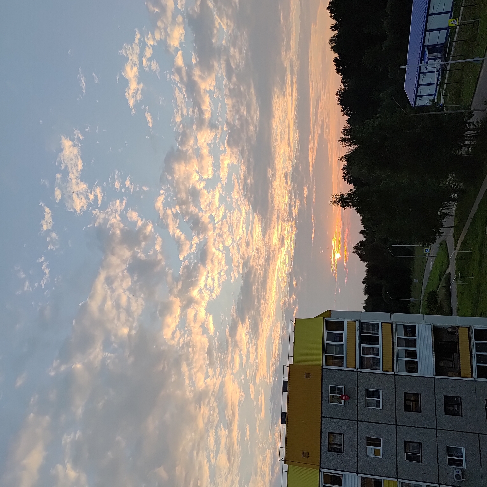 The image shows a cloudy sky at sunset over a residential area in Markovsky, Russia. In the foreground, the side of a multi-story apartment building is visible, characterized by its gray concrete facade and yellow trim on the upper floors. The building has numerous windows, some with white frames and others with darker frames, and balconies with white railings. A small red circular object, possibly a satellite dish, is attached to one of the windows.
Beyond the apartment building, a dense line of dark green trees forms the horizon. A road curves through the landscape, with streetlights and a pedestrian crossing sign visible. In the distance, to the right, a long, low building with a blue roof and white walls stands beside a chain-link fence. The sunset casts a warm, orange and pink glow on the scattered clouds, creating a serene atmosphere. The overall scene depicts a typical evening in a suburban or semi-urban environment.