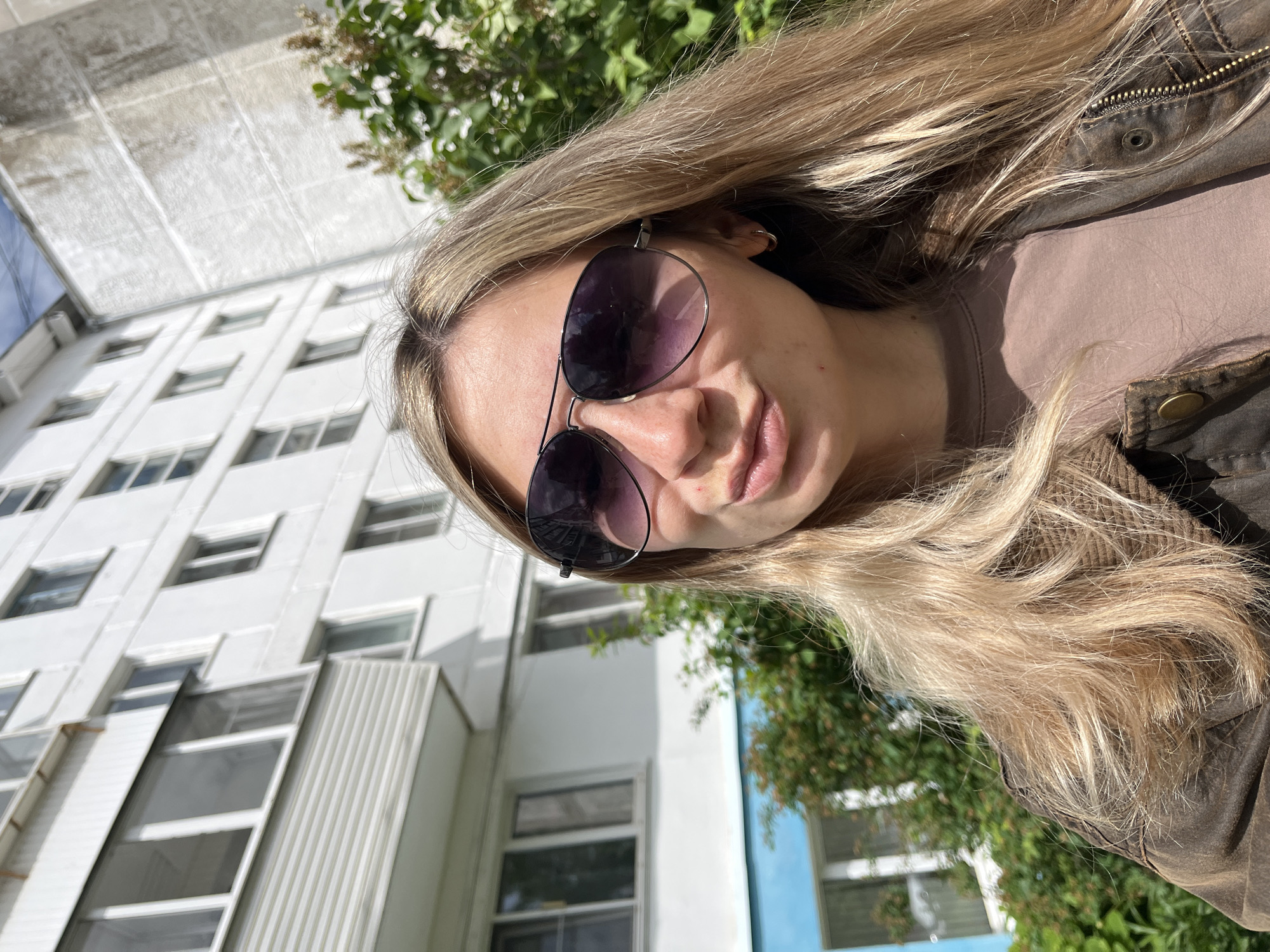 A woman with blonde hair is wearing sunglasses, a brown shirt, and a brown jacket. She is standing in front of a white building with rows of windows. Some green foliage is visible behind her. The location is in Chaikovsky, Russia.