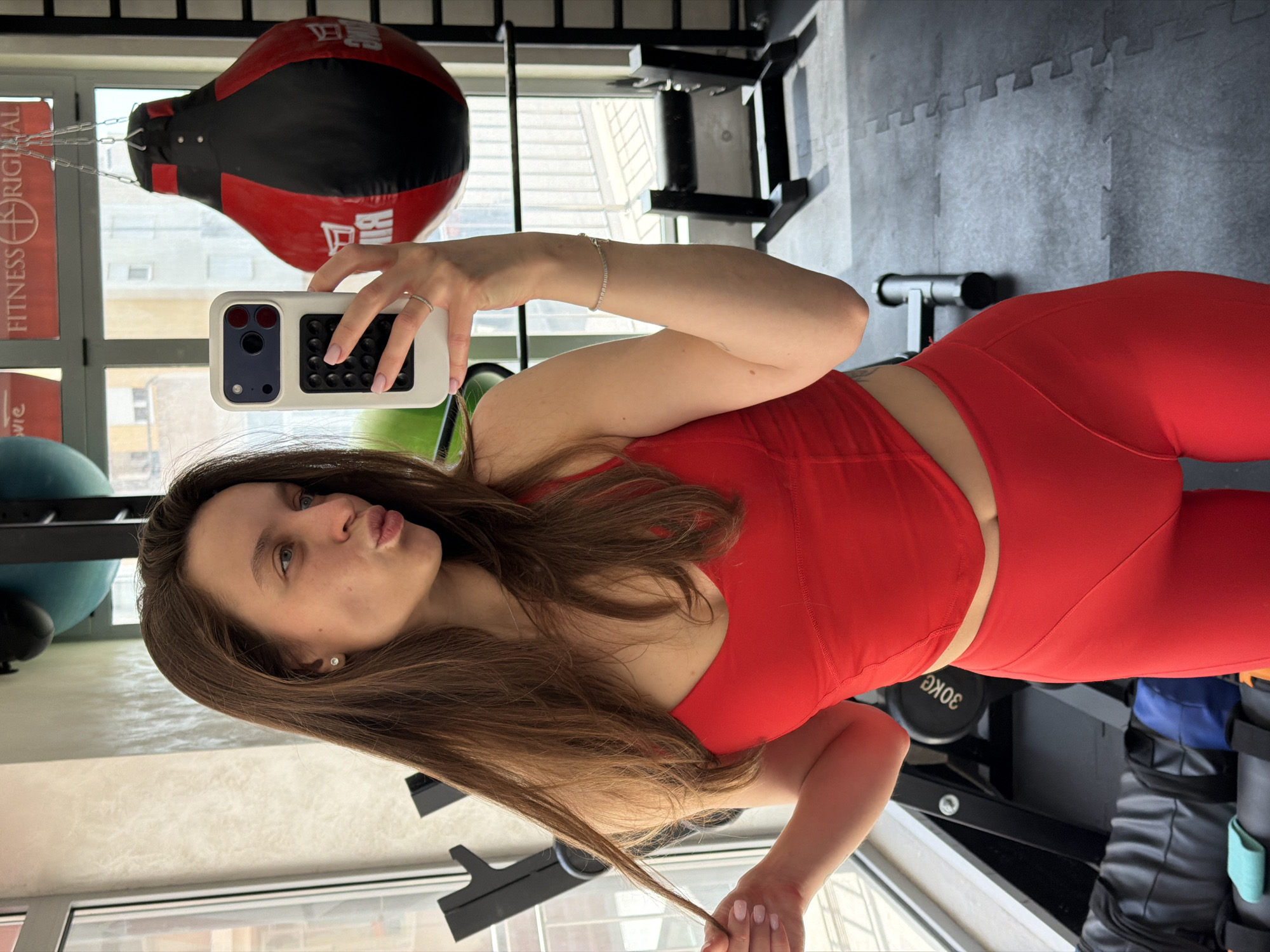 A young woman stands inside a gym taking a selfie. She is dressed in a red workout set consisting of a tank top and leggings. She holds a white phone with a patterned back, her face tilted towards the camera with pursed lips. Behind her hangs a red and black punching bag, as well as various gym equipment, including exercise balls and weight racks. The gym is located in Budva, Montenegro, as evidenced by the building visible through the window.