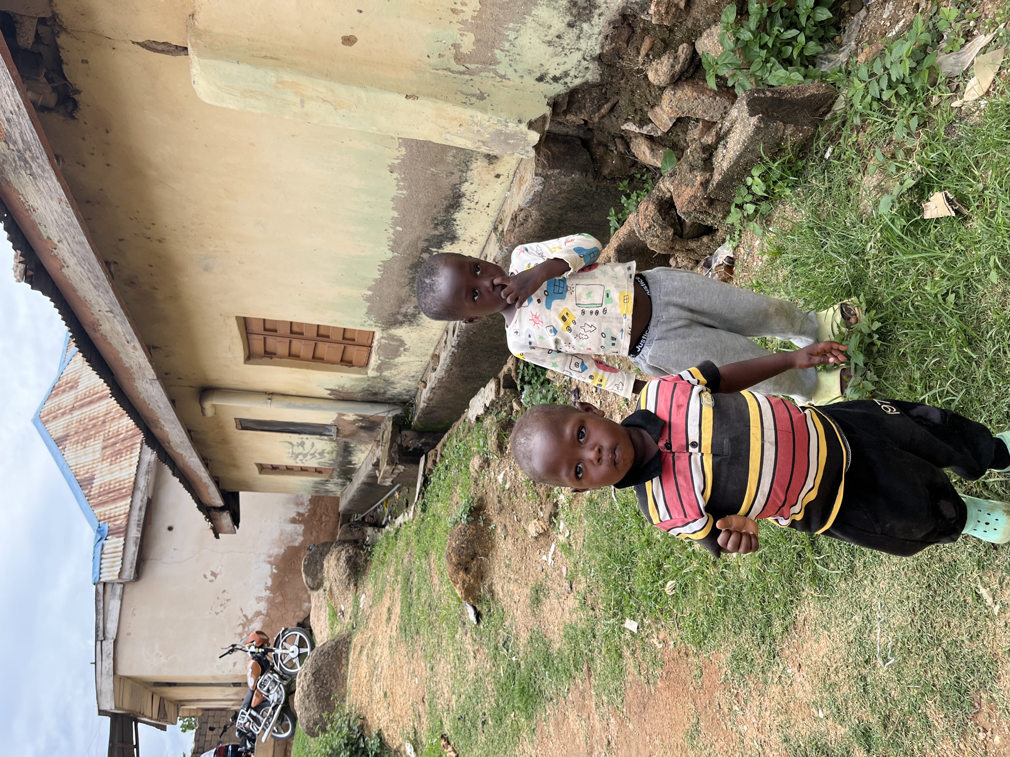 This image, captured in Jos, Nigeria, depicts an outdoor scene featuring two young children in the foreground, with residential structures and a motorcycle in the background under an overcast sky.
In the immediate foreground, a young child with dark skin and short dark hair stands facing the camera. This child wears a horizontally striped polo shirt in red, white, yellow, and black, and black trousers with the number "197" visible on the right leg. Light blue/green clogs cover their feet. Their right arm is slightly bent, and the hand is formed into a fist. To the child's right and slightly behind, a second young child, also with dark skin and short dark hair, stands looking slightly towards the left of the frame. This child wears a long-sleeved white top patterned with colorful cartoon vehicles and light grey sweatpants with "Justice" printed on the waistband. Light yellow/green sandals are on their feet. Their right hand is raised to their face, with fingers near the nose or mouth.
The ground beneath the children and extending into the mid-ground is unpaved, consisting of reddish-brown soil mixed with patches of green grass and scattered natural rocks and debris, including pieces of cardboard. A dirt path curves through the scene towards the background on the left.
In the background, two structures are visible. On the left, a light beige building with a rough-textured wall shows significant erosion or discoloration revealing reddish-brown material at its base. It has a wooden eave structure supporting a corrugated metal roof, part of which is exposed. An orange and black motorcycle with silver wheels is parked on an incline directly behind this building. On the right, a pale yellow-green building exhibits signs of weathering, including dark stains and a distinct vertical crack running down its right side. This building features a brown wooden-shuttered window and is topped with a corrugated metal roof, partially covered by blue tarpaulin in one section. A concrete drainage channel runs along the base of this building, containing dark liquid and debris. The sky above is uniformly grey and cloudy.