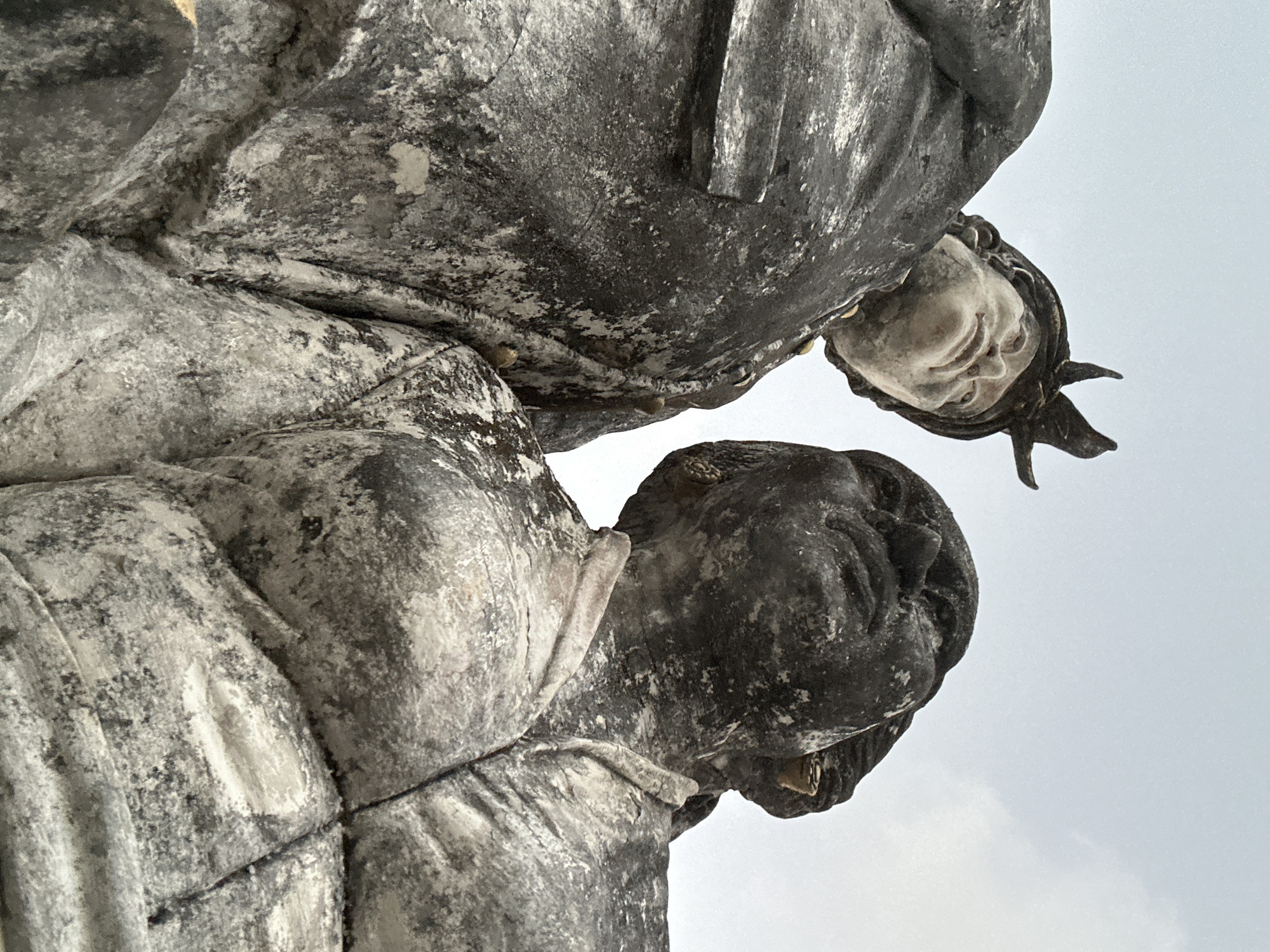 This is a low-angle, close-up, exterior shot of two weathered, stone statues in Kuta Selatan, Indonesia. The statue on the left depicts a figure wearing a helmet with upward-pointing protrusions and appears to be looking upwards and slightly to the left. The statue on the right shows a figure with a more weathered, darker complexion, also looking upwards. Both statues are heavily textured and appear to be made of stone or concrete, with significant weathering and discoloration visible, suggesting they are old. The background is a pale, overcast sky with faint clouds. The statues are positioned close together, with their upper torsos and heads filling most of the frame.
