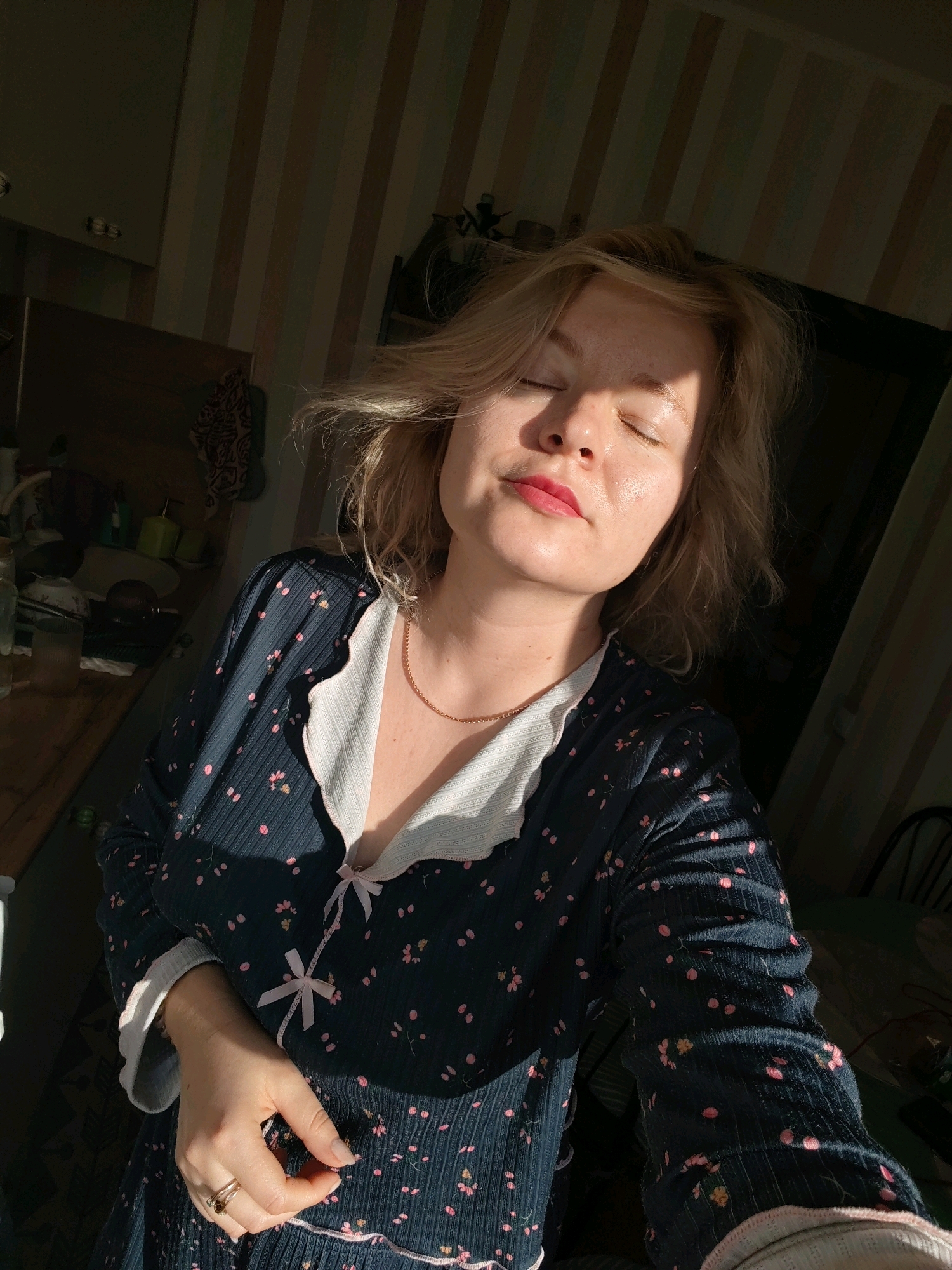 A blonde woman is indoors, with her eyes closed and face tilted up towards a light source casting shadows on her face and the striped wallpaper behind her. She is wearing a dark blue nightgown with a floral pattern of pink and yellow flowers, and a white ruffled collar. A delicate gold chain necklace is visible around her neck. Her right hand rests on her chest, and she is wearing two rings on her ring finger. The background appears to be a domestic setting with shelves and items visible in the dimmer areas of the room. The lighting suggests it is either sunrise or sunset, with strong directional light creating a chiaroscuro effect. The location is identified as Izhevsk, Russia.