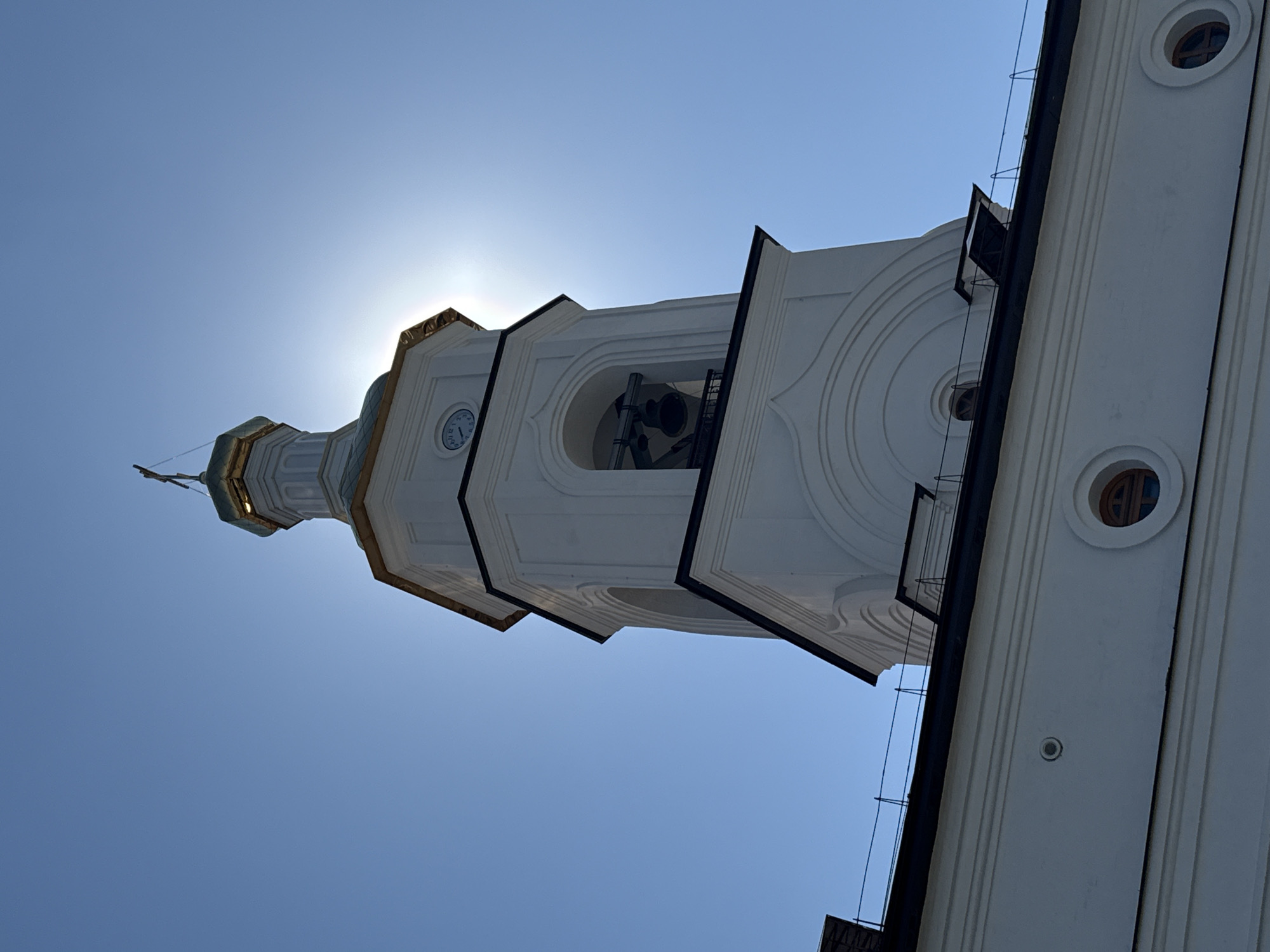 The image shows the bell tower of the Tobolsk Kremlin in Tobolsk, Russia, taken from a low angle looking upwards. The white tower is multi-tiered and features a clock face, an open bell chamber, and rounded windows. The sun is positioned behind the tower, creating a bright glow. The sky is clear and blue. A portion of an adjacent building is visible on the right side of the frame.