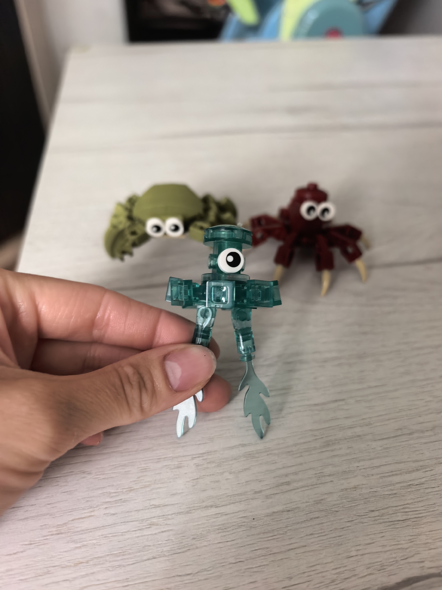 A human hand is prominently featured in the foreground, holding a translucent, teal-colored toy figure. This figure, composed of various connected block-like pieces, features a single large googly eye and two light-blue, leaf-shaped appendages at its base. The hand, with visible skin texture and fingernails, holds the figure upright.
In the blurred background, two other whimsical toy figures rest on a light-colored wooden surface with a distinct grain pattern. To the left, an out-of-focus green, crab-like figure with multiple legs and two googly eyes is visible. To its right, a reddish-brown, spider-like figure with several pointed legs and two googly eyes can be seen. All the toys share a playful aesthetic, primarily due to their googly eyes.
The scene takes place indoors, likely on a table or desk. The activity depicted is the holding and presenting of one of these unique toy figures, possibly for play or display. There is no visible text in the image. The time of day or weather cannot be determined. The provided location context is Sterlitamak, Russia.