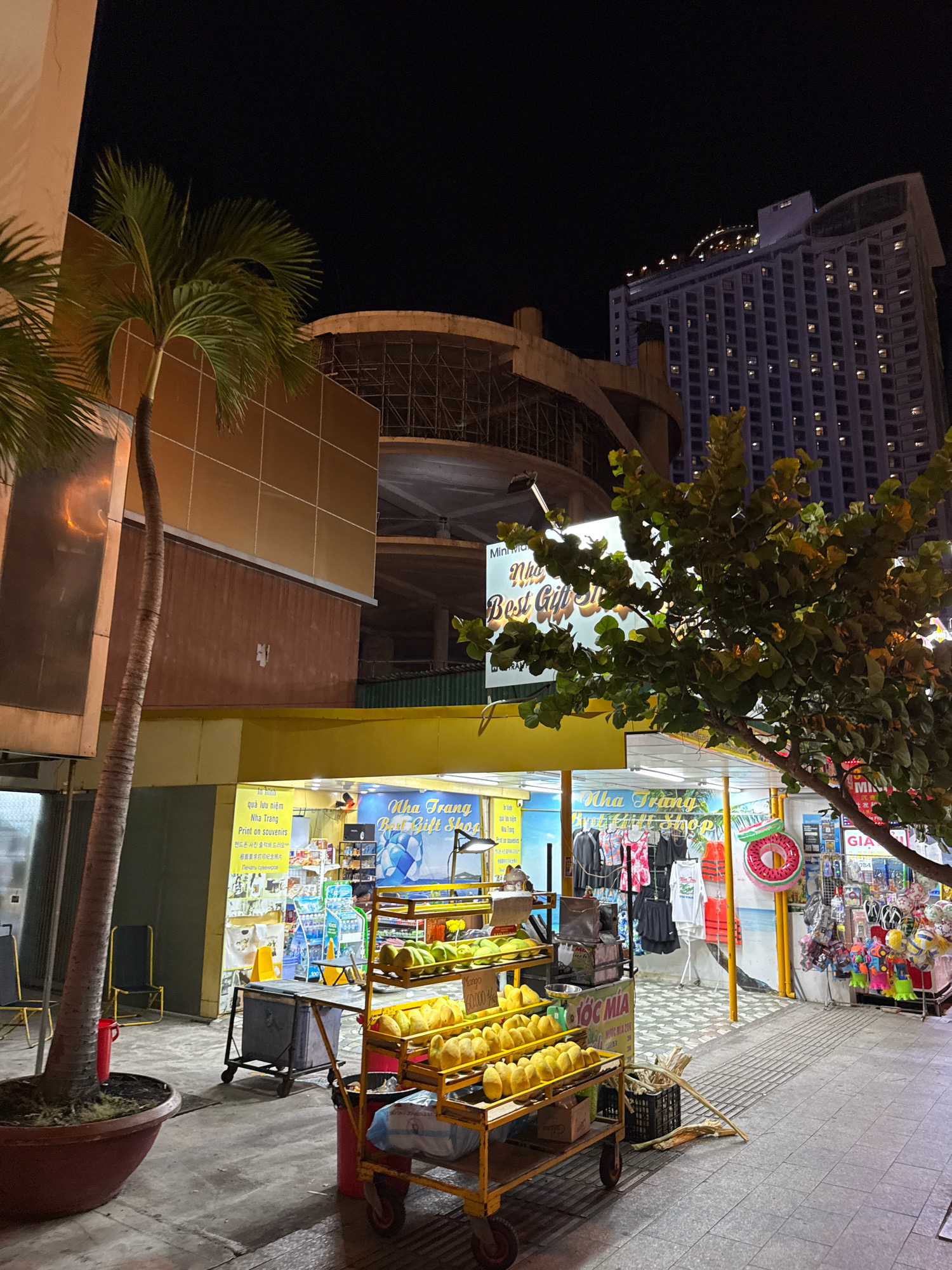 The image depicts a street scene at night in Nha Trang, Vietnam, specifically in front of a store and food stalls. A prominent sign above the store reads "Nha Trang Best Gift Shop" in English and Vietnamese. The store appears to sell various items including clothing, souvenirs, and possibly food. To the left of the store, there are shelves displaying what seem to be tropical fruits, likely mangoes, on a yellow cart. A person, partially visible, is working behind a counter inside the store.
In the background, a tall, modern building with many illuminated windows rises into the dark night sky, characteristic of a city skyline. To the left of the building, there's a partially constructed structure with scaffolding, suggesting ongoing construction. In the foreground, a large potted palm tree stands to the left, and a leafy tree partially obscures the view of the store and the sky. The ground is paved with tiles and a concrete sidewalk. The overall scene suggests a lively commercial area in Nha Trang, with shops and food vendors operating into the evening.
