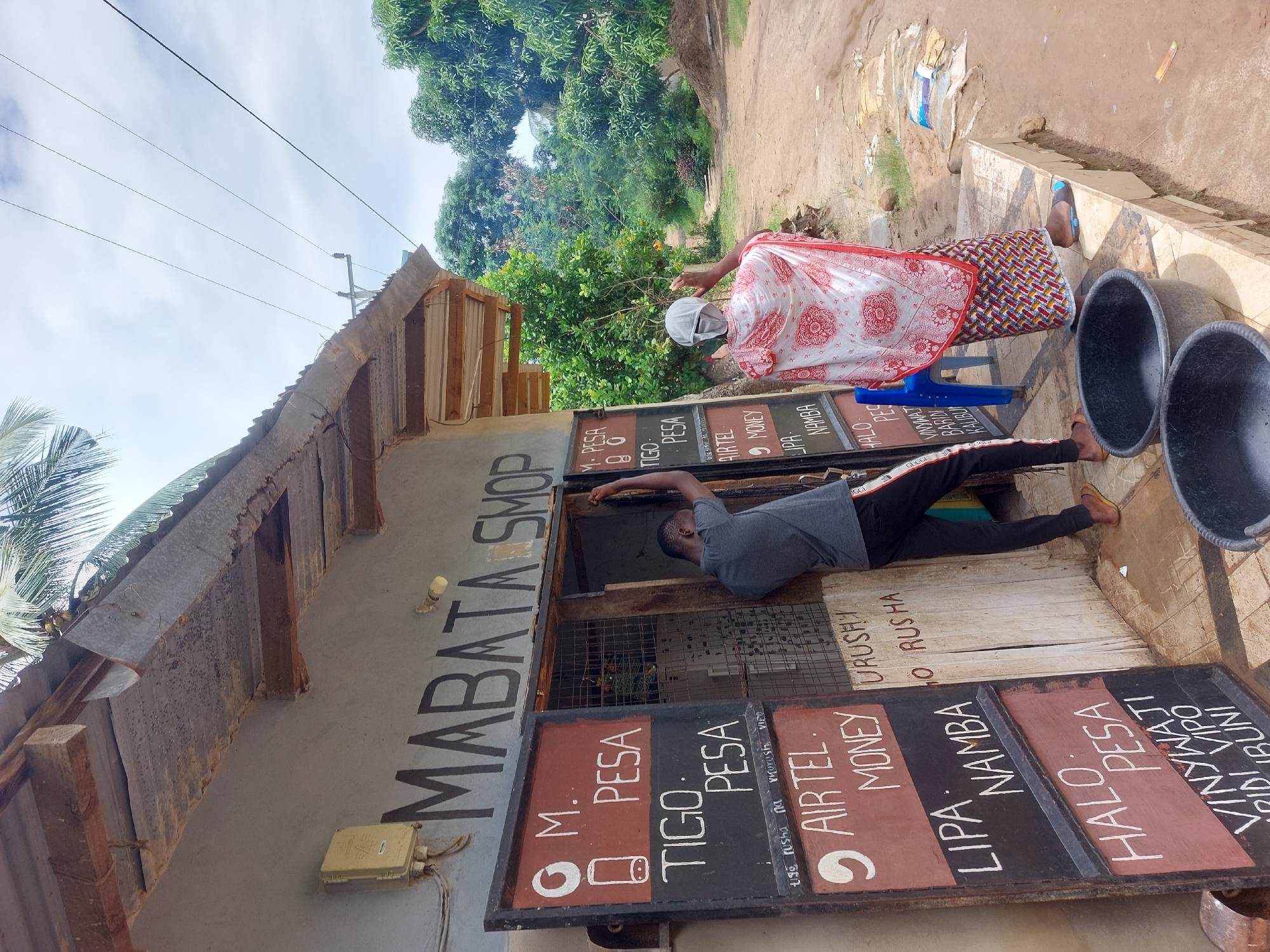 The image depicts a scene at a small shop called "MABATA SHOP," likely located in Tanzania, given the Swahili language used ("pesa" means money). The shop appears to offer mobile money services ("M-PESA," "TIGO PESA," "AIRTEL MONEY") and possibly sells drinks ("VINYWAJI," cold drinks "BARIDI" "KALIBU"). A man is leaning into the shop, possibly interacting with someone inside, while a woman stands outside wearing a head covering and a colorful patterned wrap. There are black plastic basins and a blue plastic chair nearby. The environment looks somewhat rural, with trees visible in the background, but a definitive location is impossible without further context. The shop itself is a simple structure with a tin roof and signage indicating the services it provides.
