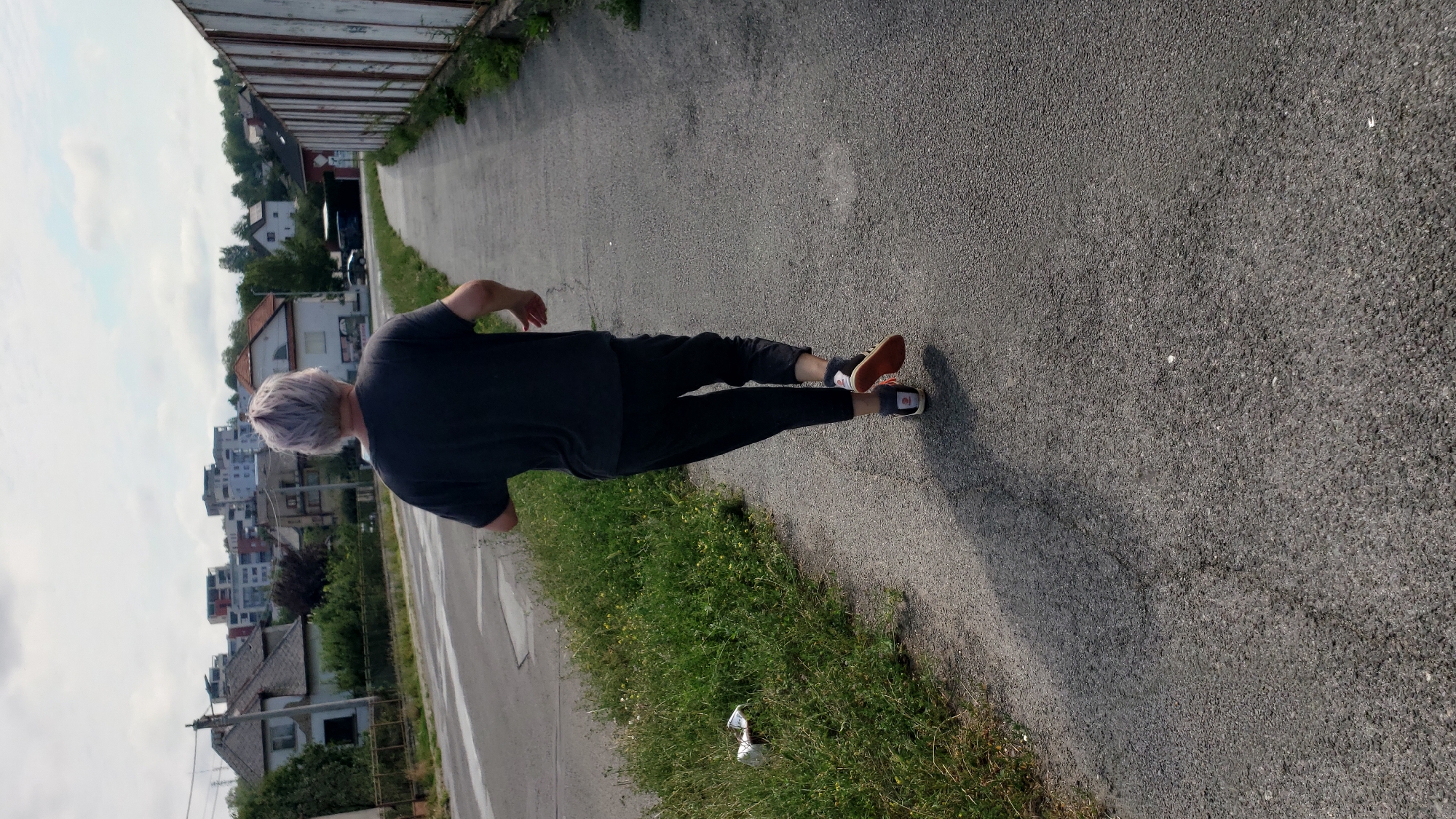 A person with gray hair is running away from the camera down a paved path in Prešov, Slovakia. They are wearing a dark t-shirt and dark pants. The path is next to a road with some grass and a few pieces of litter on the side. In the background, there are houses and apartment buildings. A tall, white fence runs along the right side of the path.
