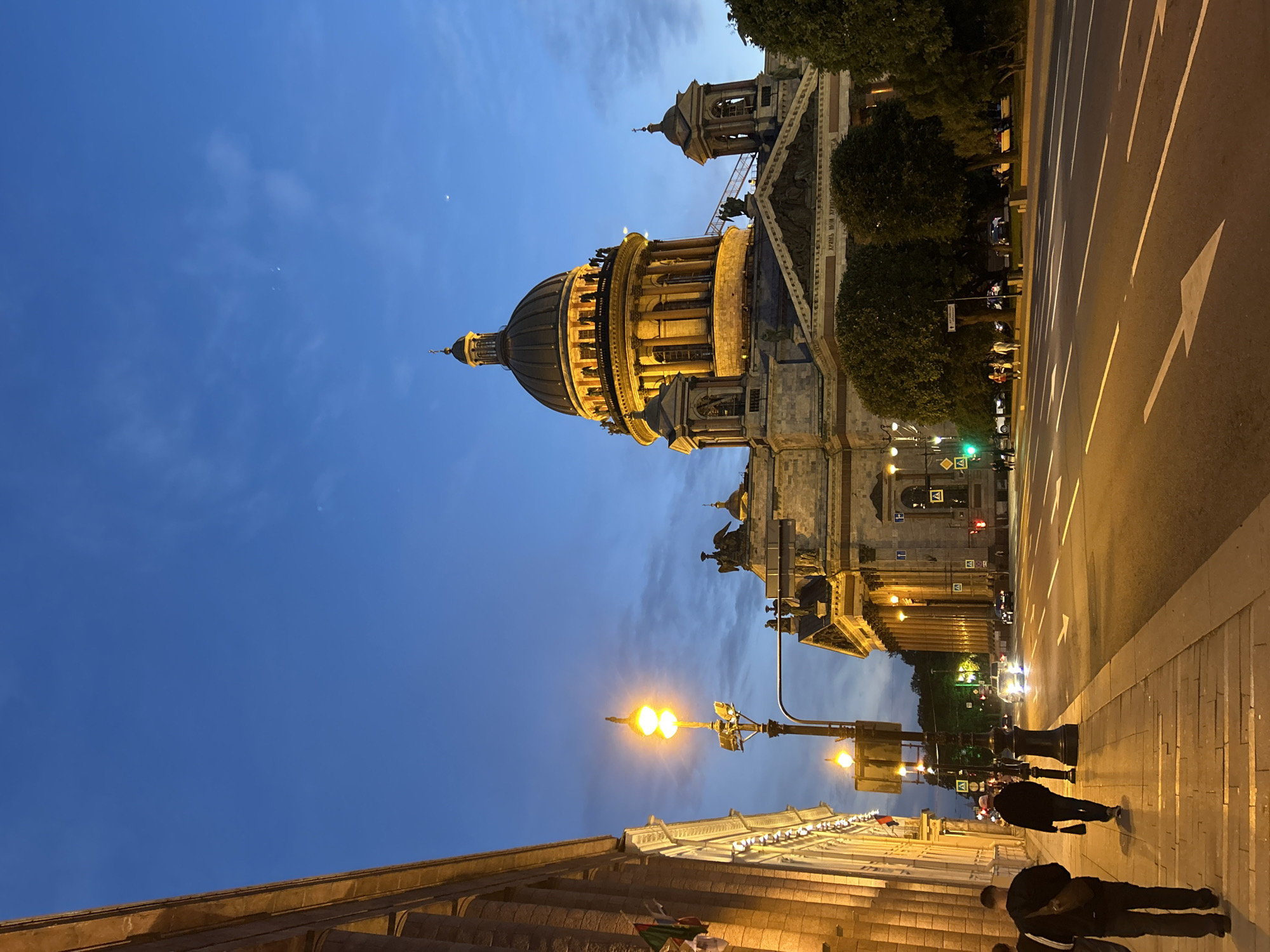 In the city of Unknown, Unknown, a majestic domed building, illuminated by warm light, dominates the scene. The sky above is a deep twilight blue, with wisps of clouds and a faint crescent moon visible. In the foreground, a street stretches out, lined with a sidewalk where several individuals walk. A streetlamp with two bright lights illuminates the sidewalk and a portion of the road. The building's facade features classical columns and ornate architectural details, with statues adorning its upper levels. Green trees are interspersed around the structure, adding a touch of nature to the urban landscape. Cars are visible on the road, suggesting the ongoing activity of the city. The overall atmosphere is one of serene evening grandeur.