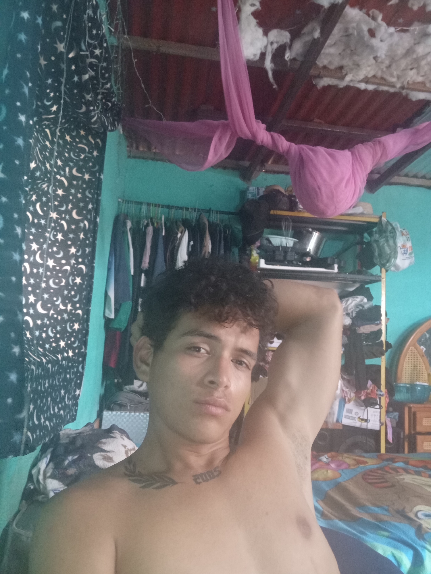 The image is a selfie taken in an indoor setting. A young man with curly dark hair is positioned in the foreground, with his right arm raised and resting behind his head. He is shirtless and has tattoos on his left shoulder and chest, including the number "2005".
The background reveals a cluttered living space. To the left, a dark curtain with a white star and moon pattern is visible. In the center, hanging clothes are organized on a rack against a turquoise wall. Above the clothes rack, shelves are stacked with various items, including kitchenware, bags, and what appears to be bedding. A pink mosquito net or fabric is draped across the ceiling, which is made of corrugated metal. The overall environment suggests a modest, lived-in home.
The location is identified as Altagracia, Venezuela. The image captures a personal moment within this specific geographical context.