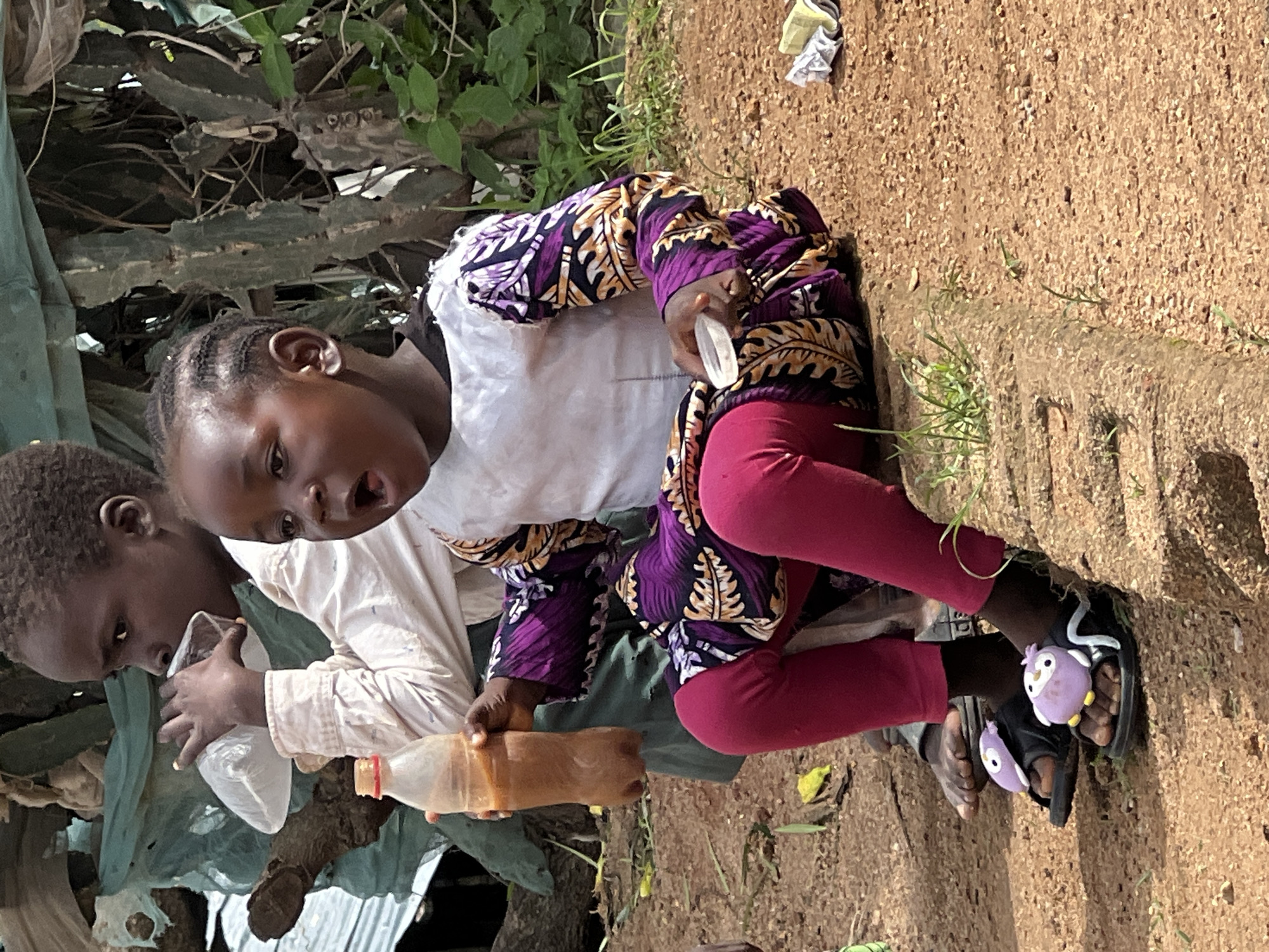 In an outdoor environment on the outskirts of Jos, Nigeria, a young Black child is seated on a raised dirt embankment. The child is wearing a white top with purple and gold patterned sleeves and leggings. They are holding a clear plastic bottle with a brown liquid and a small white lid. To the left, another child, partially visible, is drinking from a clear plastic bag. The background consists of lush green vegetation and trees. The ground is covered in reddish-brown dirt and scattered green grass. The child's feet are adorned with black sandals featuring purple owl-shaped decorations.