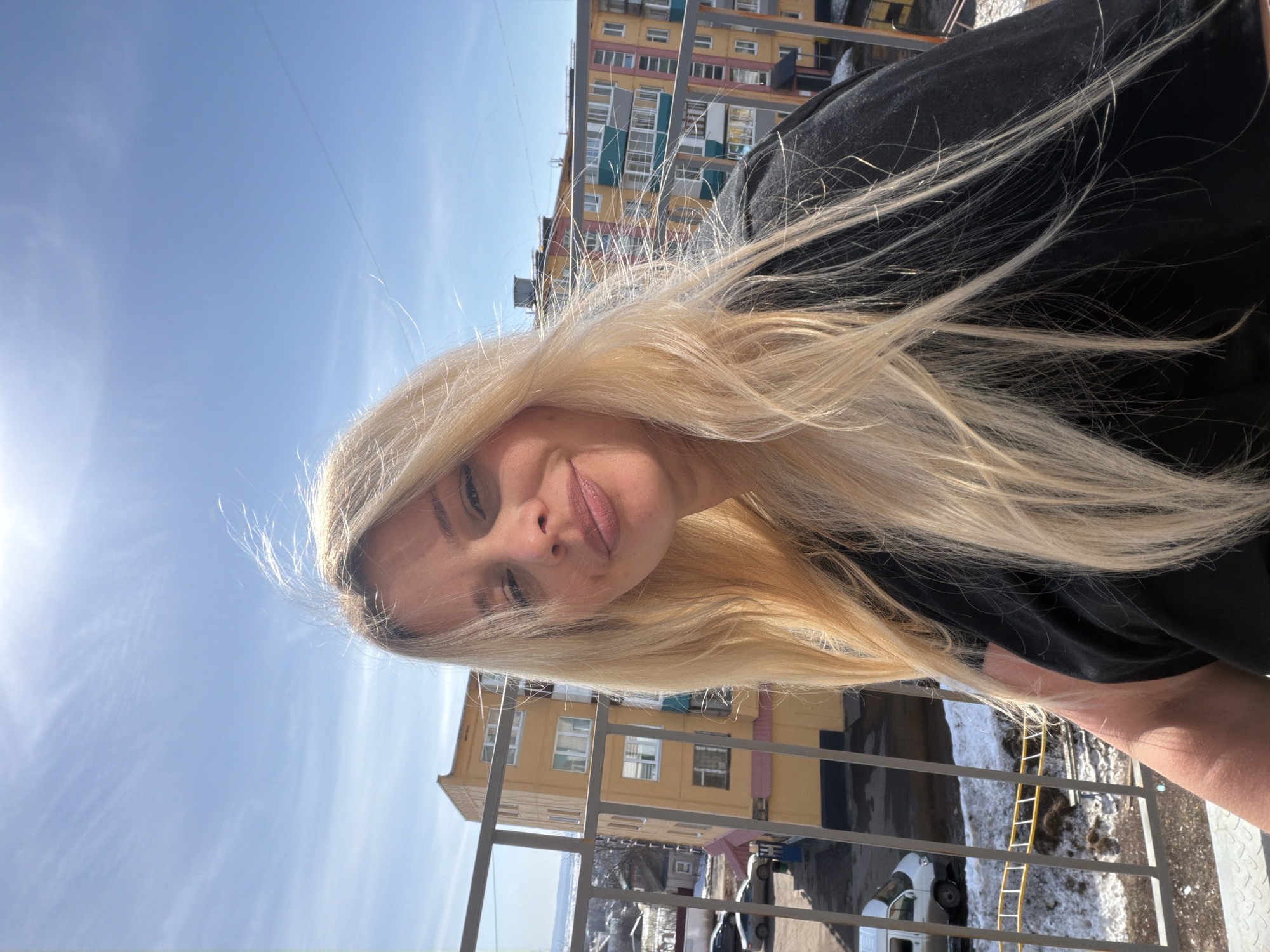 A selfie of a blonde woman in Sarapul, Russia. She is wearing a black shirt and has long blonde hair. She is standing on a balcony with a view of a building and a blue sky. A yellow ladder lies on the ground outside of the railing.