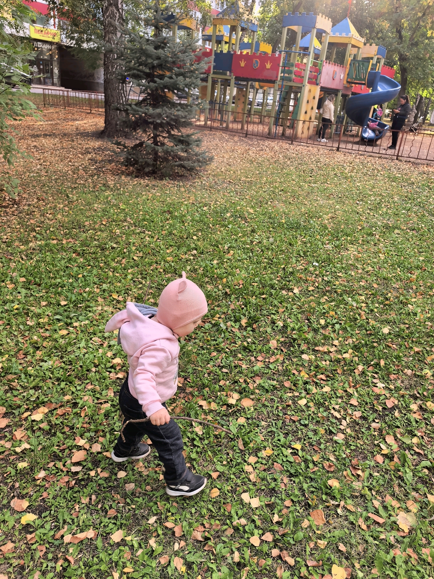 A toddler, wearing a pink hooded sweatshirt with ear details on the hood and dark pants, walks across a lawn covered in fallen leaves. The child is holding a stick. In the background, a brightly colored playground with slides and climbing structures is visible. Several adults are near the playground. The scene appears to be a park or public space in the city of Sterlitamak, Russia, suggested by the presence of a playground and a building with Cyrillic signage that appears to be a bakery (indicated by "ПЕКАРН" visible on the signage). The overall environment suggests an autumn day.