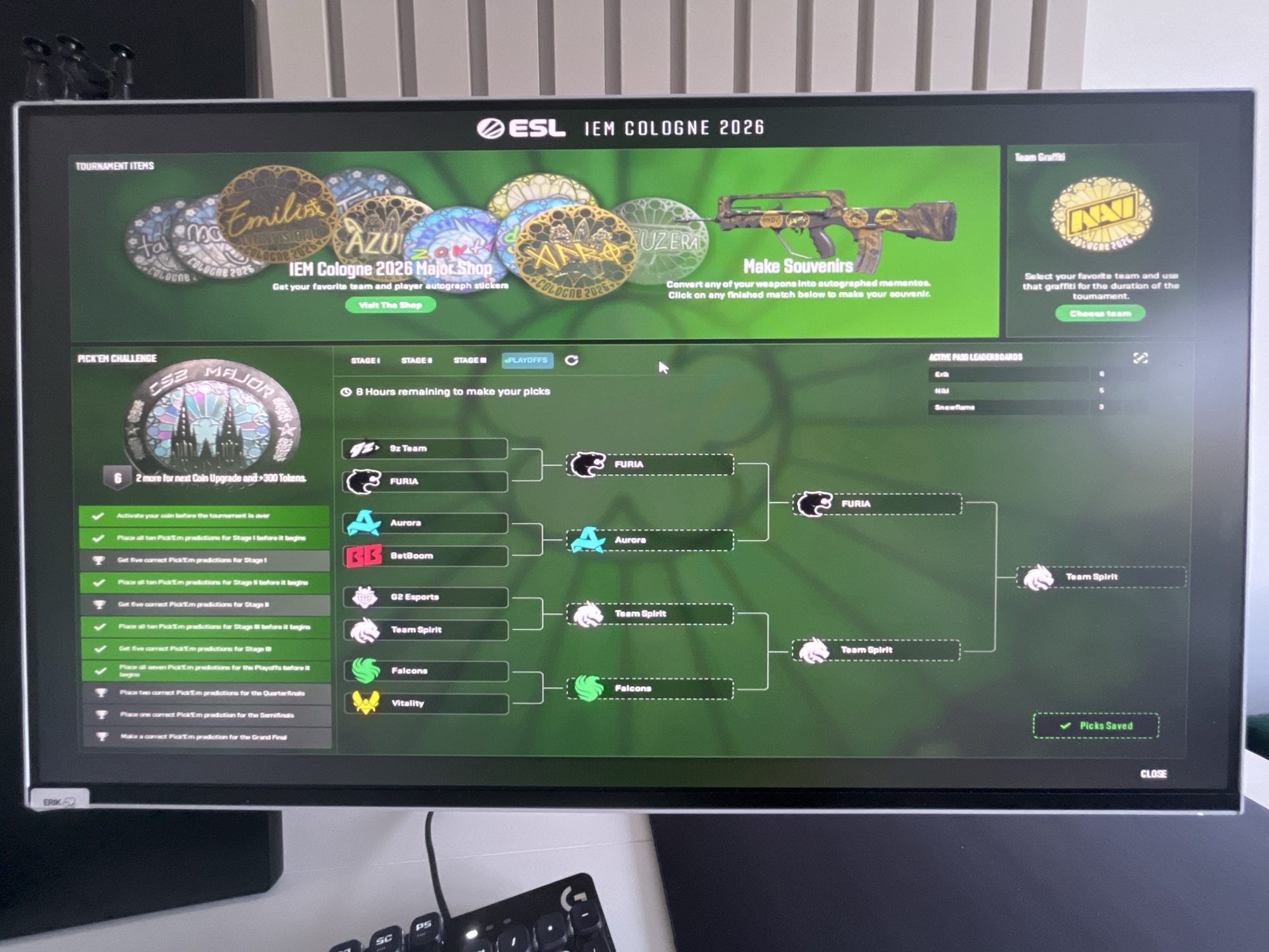 The image shows a computer monitor displaying the ESL IEM Cologne 2026 esports tournament bracket. The screen shows a "Pick'Em Challenge" with several teams listed in a tournament bracket format. The tournament is for CS2 (Counter-Strike 2). The screen also displays tournament items, including collectible stickers and weapon skins. The "Team Graffiti" section shows the Natus Vincere (Na'Vi) team logo, suggesting it's a choice for team-specific in-game cosmetic items. The bracket shows matches for various stages of the tournament, with some picks already made and indicated as saved. The environment is consistent with a home office or gaming setup, with part of a keyboard visible in the foreground. There are no indications of the city of Varna, Bulgaria, in the image.