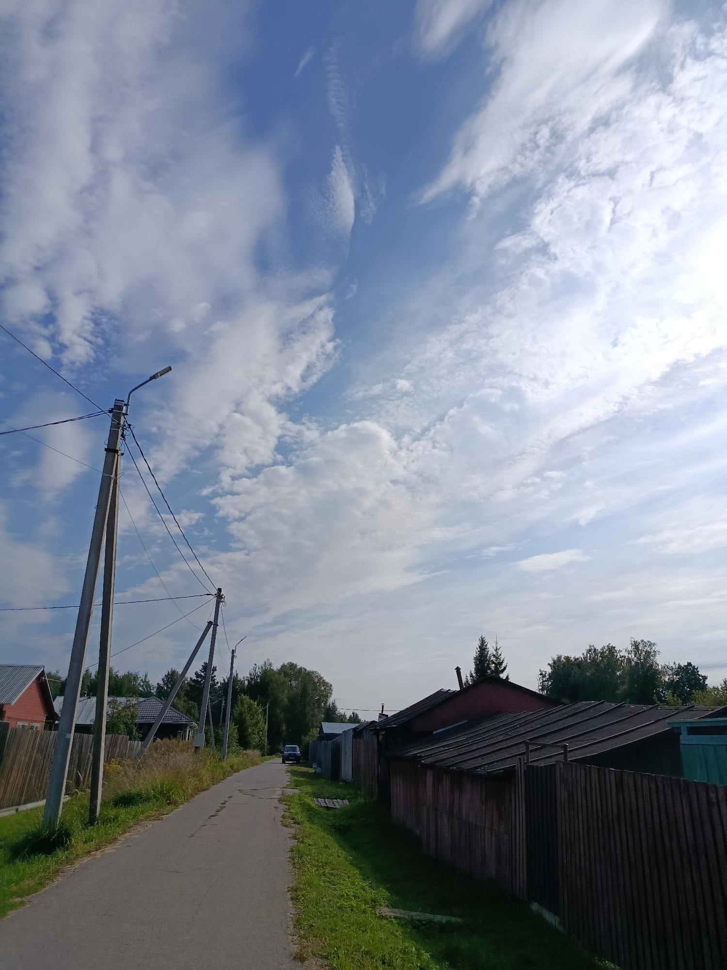 This is a street view photograph taken in the city of Vereya, Russia. The image shows a paved road leading into the distance, flanked by grassy verges. On the left side of the road, there are houses with wooden fences and tall concrete utility poles with electrical wires. On the right side of the road, there are buildings with dark roofs and wooden fences. Further down the road, a dark-colored SUV is visible. The sky is blue with scattered white clouds. The overall environment appears to be a rural or suburban residential area.