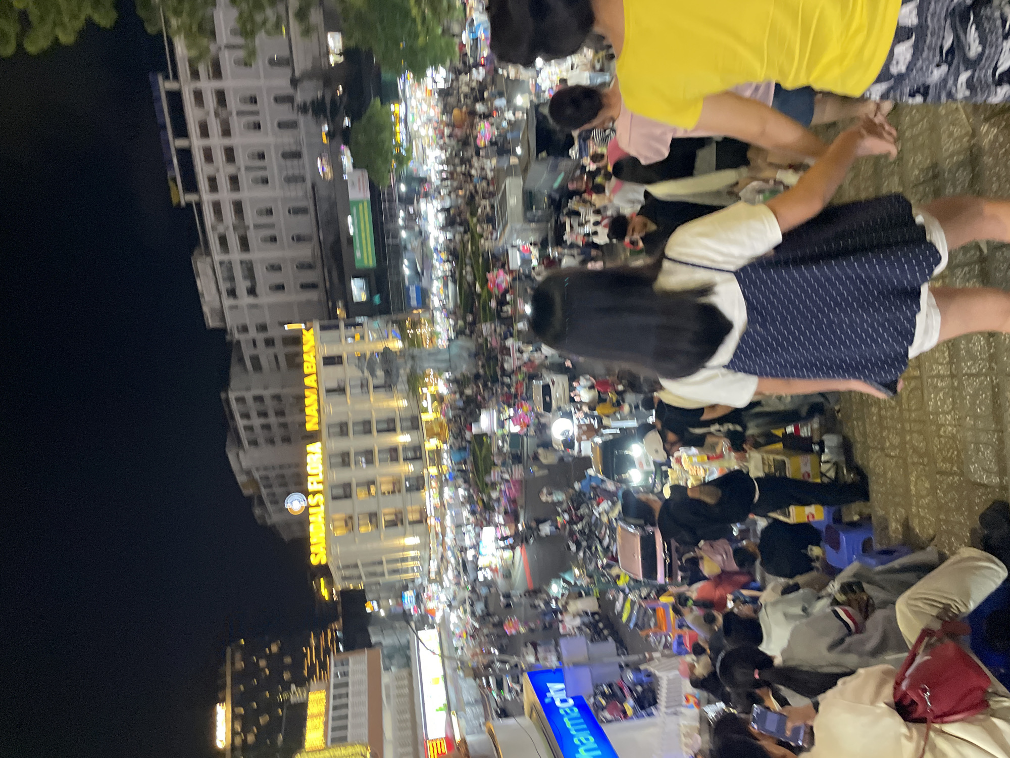 The image captures a bustling night scene in Da Lat, Vietnam, likely a market or busy street area. The foreground shows a group of people walking down stone steps. In the midground, a dense crowd navigates a street filled with vendors, motorcycles, and cars. Buildings with illuminated signs, including "Sandals Flora Namabk" and "pharmacity," line the street, indicating a commercial district. The overall impression is one of vibrant nightlife and activity.
