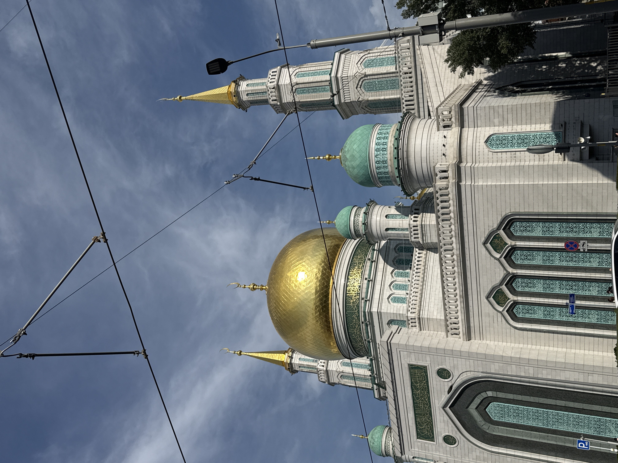 The image shows a close-up, low-angle view of the Moscow Cathedral Mosque, a prominent Islamic landmark in Moscow, Russia. The mosque is characterized by its grand white marble facade, intricate architectural details, and multiple domes and minarets.
The central and largest dome is a brilliant gold, reflecting the sunlight. Two tall, slender minarets with gold-tipped spires flank the main dome, and smaller green-domed structures are visible on either side. The building's exterior is adorned with decorative elements, including patterned windows and Arabic calligraphy in green script above some of the arched entrances.
In the foreground, overhead power or trolley wires crisscross the sky, obscuring parts of the upper structures. A streetlamp with a modern LED fixture is visible on the right side of the image, attached to a pole that also supports several electrical wires. In the lower left corner, a blue parking sign with a white "P" is visible. Further to the right, traffic signs are present, including a red and blue circular sign. A car is partially visible at the bottom of the frame. The sky above is a mix of blue with scattered white clouds, indicating daytime. The lighting suggests it is daytime, possibly late morning or afternoon, with the sun casting shadows on the mosque's facade. No people are clearly discernible in the image, though a few figures might be present near the bottom.