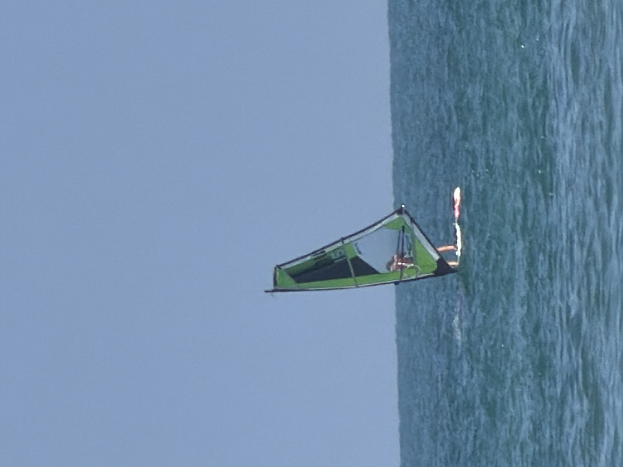 A windsurfer is sailing on the Mediterranean Sea off the coast of Tel Aviv, Israel. The sky is clear and blue. The water is choppy. The windsurfer has a green and black sail. The windsurfer is wearing a life vest.