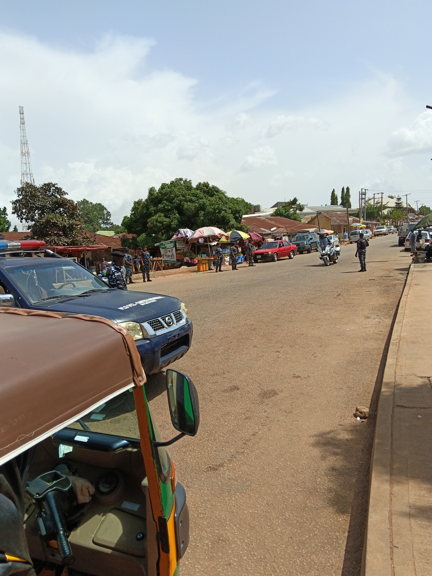 The image depicts a street scene in an outdoor marketplace, likely during daytime under a cloudy sky. In the foreground, the interior of a brown and yellow auto-rickshaw is visible, with its steering wheel and passenger seats. Beyond the rickshaw, a blue police SUV is parked, with its emergency lights illuminated on the roof. Several police officers in uniform are standing near the SUV and in the street.
The background shows a street lined with market stalls and various vehicles, including cars, another police motorcycle, and more auto-rickshaws. Some of the stalls appear to be selling fruits and other goods, with umbrellas providing shade. The overall atmosphere suggests a busy commercial area, possibly with some form of security presence or event taking place due to the visible police activity. The architecture of the buildings and the style of the vehicles do not provide immediate clues to a specific location, but the scene is consistent with many urban or semi-urban environments. The time of day appears to be late morning or afternoon, based on the lighting.