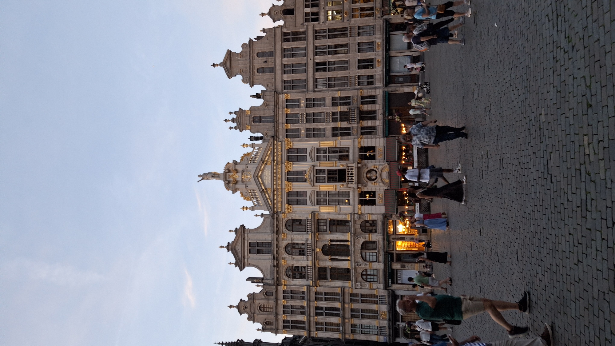 The image captures a wide shot of a European city square, likely during dusk, with an ornate, gilded building dominating the background. The building features multiple stories with arched windows and decorative elements. A statue stands atop the central section of the building. The square is paved with cobblestones, and several people are visible walking around. Some are dressed casually, while others appear to be tourists. The sky is a pale blue with some wispy clouds. The lighting suggests a warm, evening atmosphere.