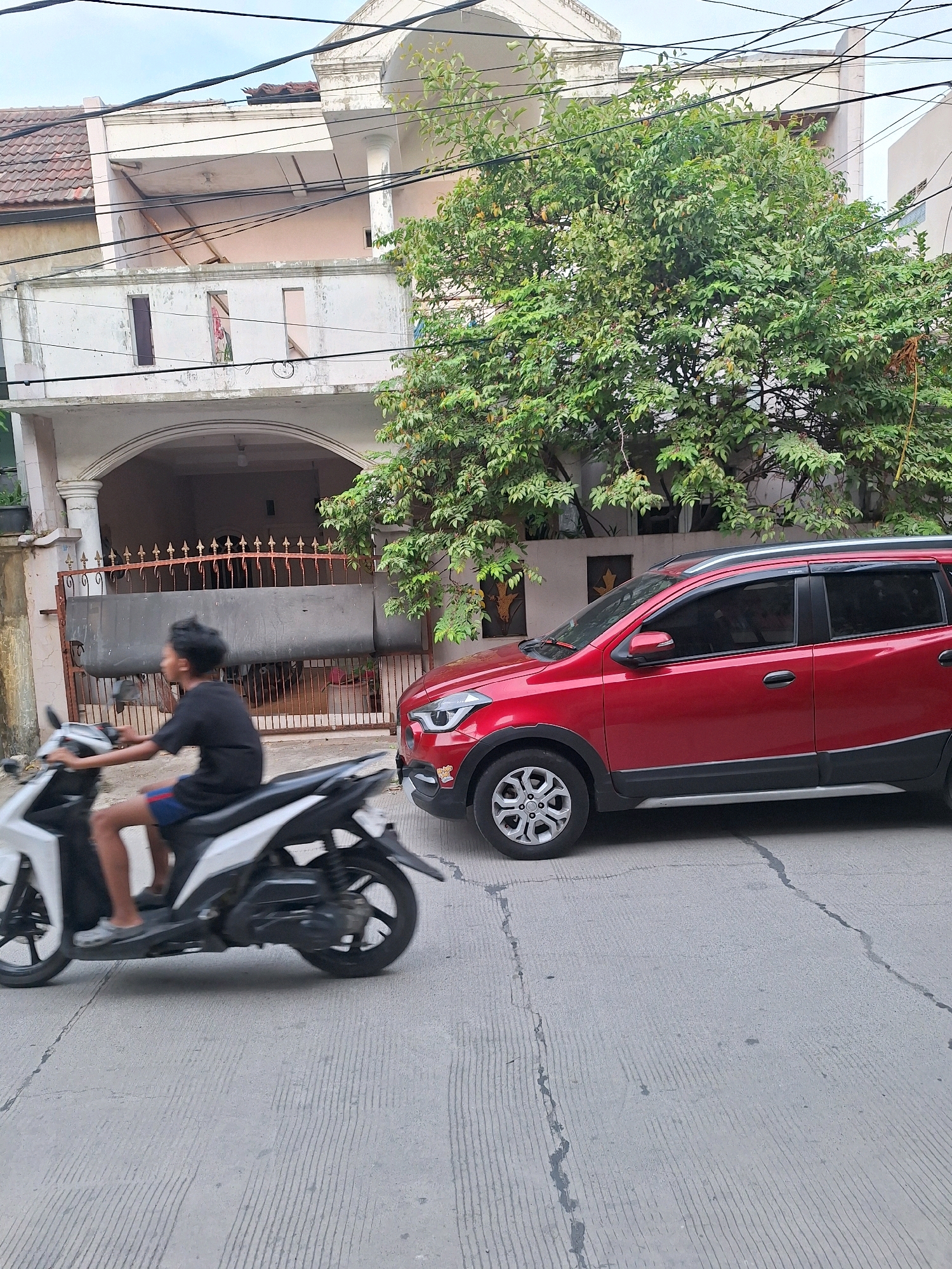 A young person rides a white scooter down a paved street in Setiamulya, Indonesia. The rider is wearing a black t-shirt and blue and red shorts. A red SUV is parked on the right side of the street. In the background, there is a multi-story house with an arched entryway and an ornate metal gate. A large tree with green leaves is in front of the house. The sky is overcast.