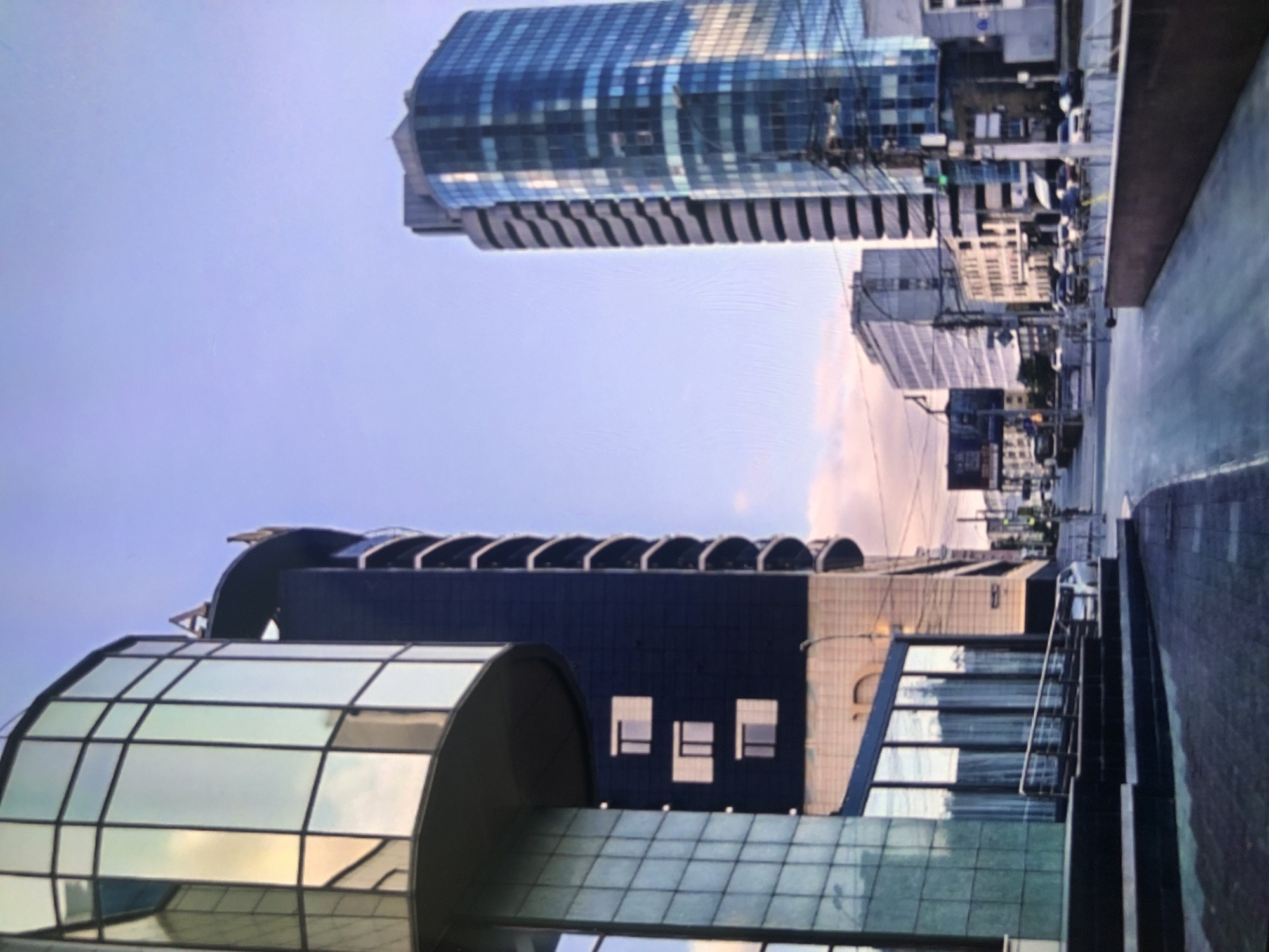 The image presents an urban scene in an unidentified city. The perspective is from ground level, looking upward towards a complex of modern buildings. The foreground is dominated by a structure featuring curved glass panels and dark structural elements. Behind this building, a taller structure with a dark façade and a series of arched elements is visible. In the distance, a skyscraper with a grid-like glass exterior rises into a pale sky. The street appears to be relatively empty, with only hints of traffic and street furniture. The scene lacks people and any indications of specific actions or interactions.