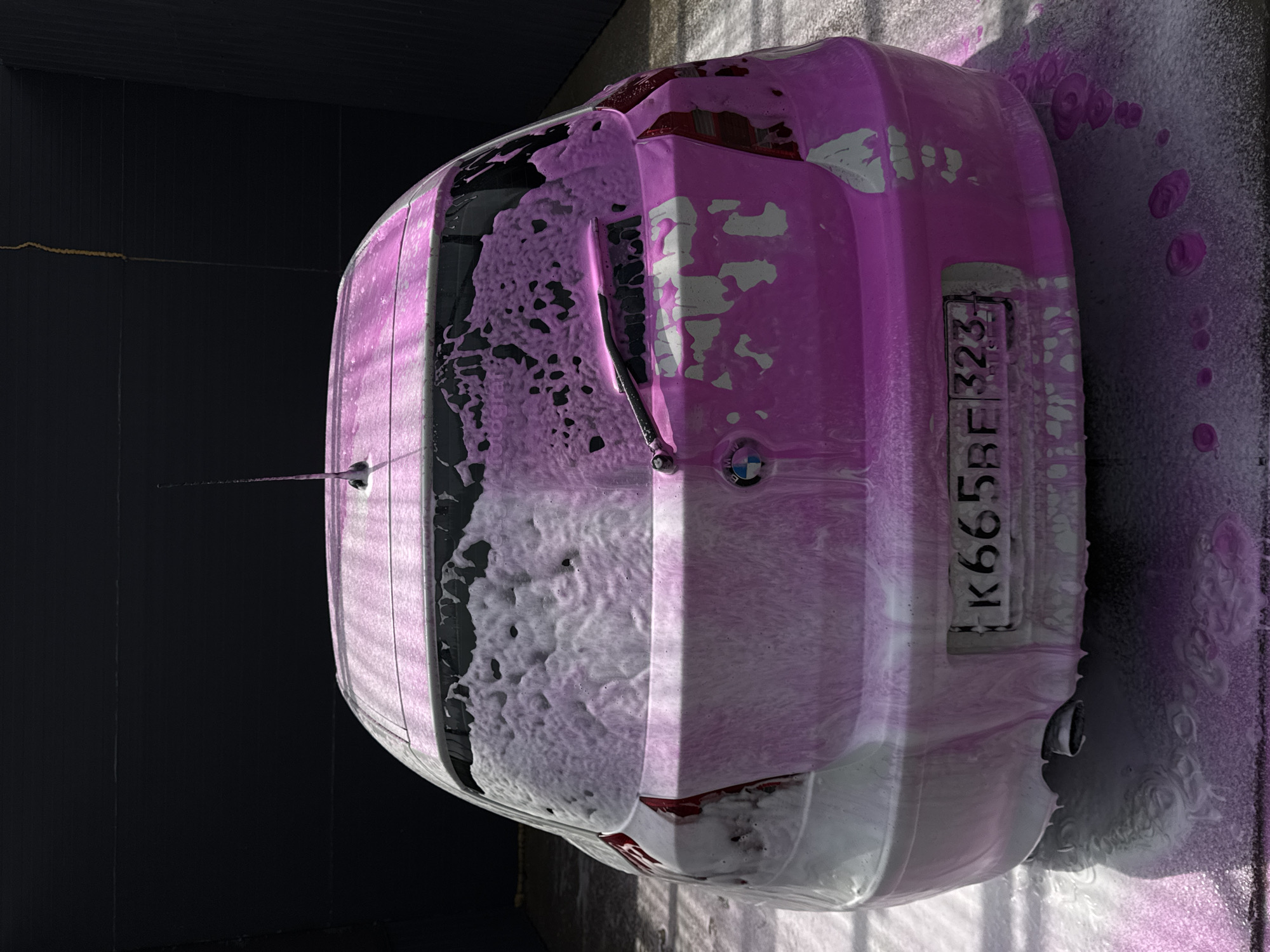 A BMW car with a Russian license plate number K665BE 323 is covered in pink foam, presumably during a car wash. The car is indoors with a black wall visible behind the rear window. The car is tilted to the right. The location is likely in or near Rossiyskiy, Russia, based on the license plate format.