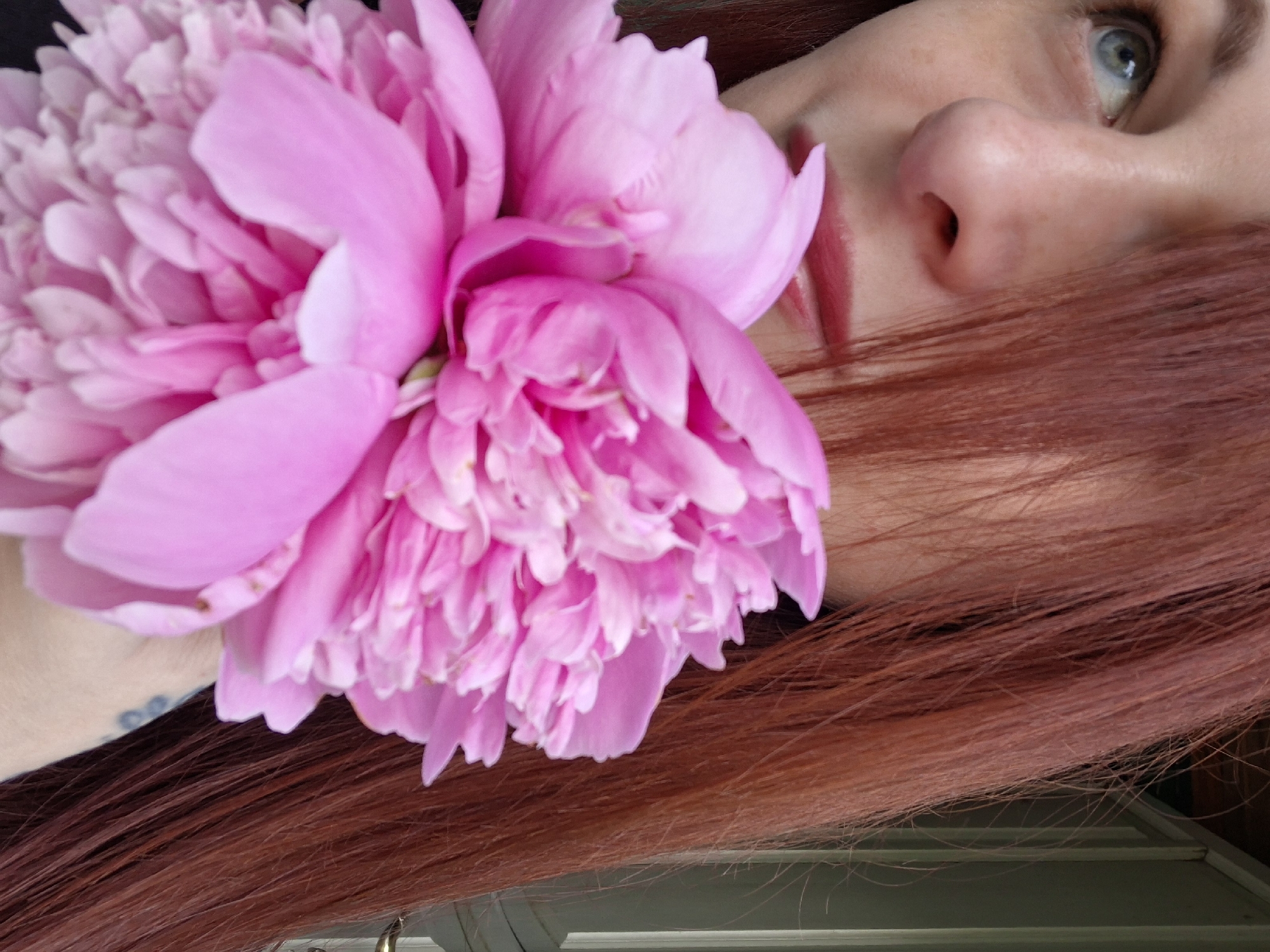A close-up portrait shows a woman with long, reddish-brown hair and light blue eyes. She is holding a large, pink peony flower in front of her face, obscuring her nose and mouth. Her lips are painted with a reddish lipstick. The background is out of focus, but appears to be an outdoor setting with a green door or wall visible on the right. The lighting suggests it is daytime. No specific location in Забалоцкі сельскі Савет, Belarus can be identified from the image.