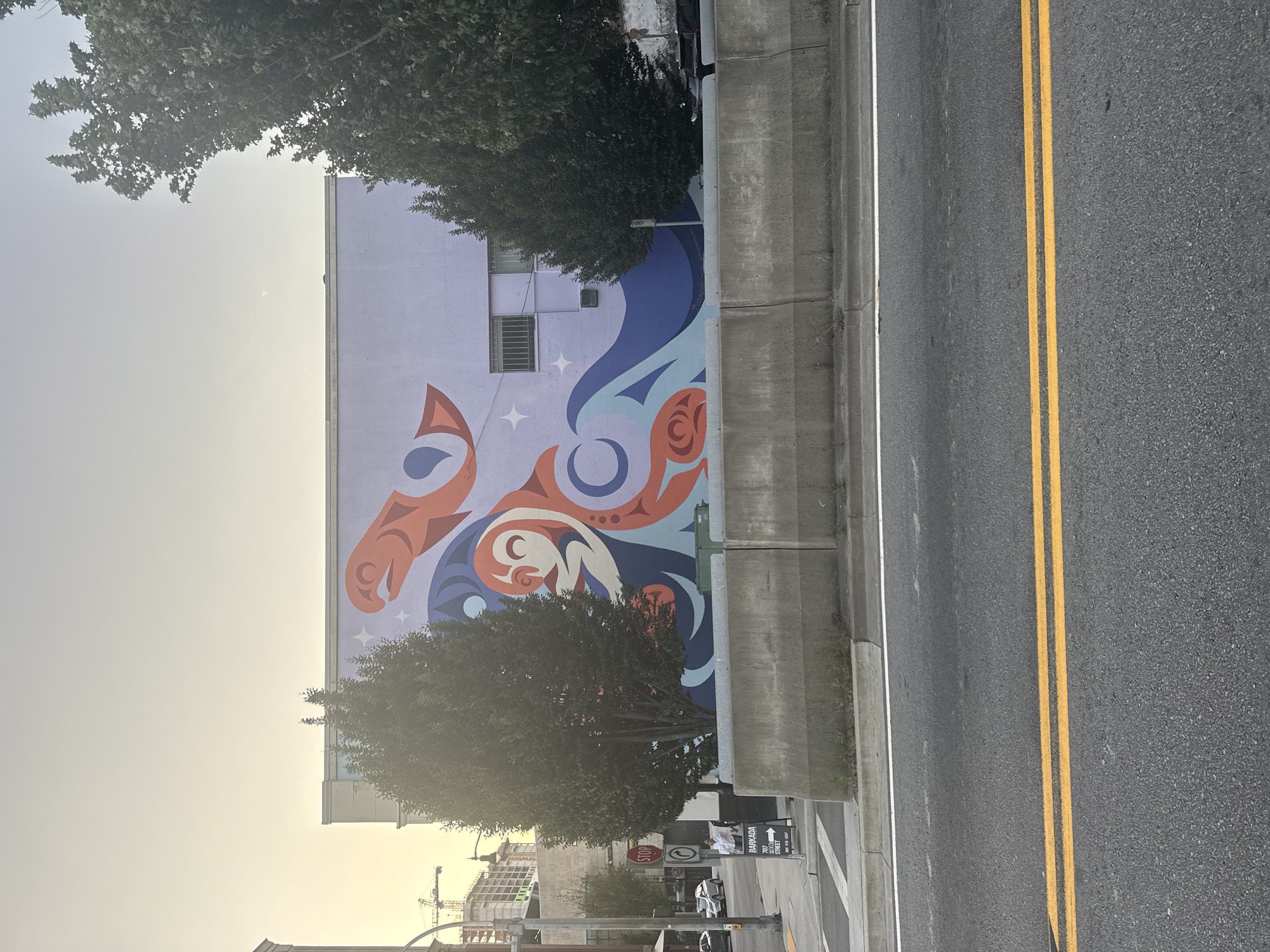 The image captures a wide shot of a mural on the side of a building, located in New Westminster, Canada. The mural, painted in shades of purple, orange, and blue, depicts stylized oceanic imagery, including waves, a fish-like creature, and a crescent moon. The building itself is a pale purple with some windows that have metal bars. In front of the building, a concrete barrier runs horizontally across the frame, separating the mural from the street. Two trees, one on the left and one on the right, partially obscure the mural. On the sidewalk, a person in a white shirt stands near a sign for "BARKADA" with an address and phone number. Cars are visible on the street in the background, along with construction cranes and other buildings, suggesting an urban environment. The sky above is a pale, hazy yellow, indicative of either early morning or late afternoon light. The foreground shows a paved road with double yellow lines.