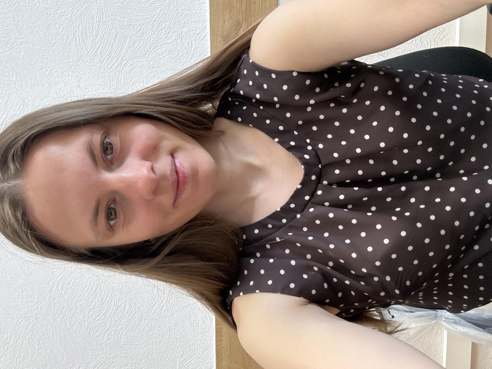 A woman with light brown hair and hazel eyes is wearing a dark brown polka dot blouse. She is indoors, with a textured white wall behind her to the left, and a light-colored wooden surface above her and to the right. She appears to be seated, with black material visible behind her torso. The location is unknown, as the image provides no information connecting it to the city of Chaikovsky, Russia.