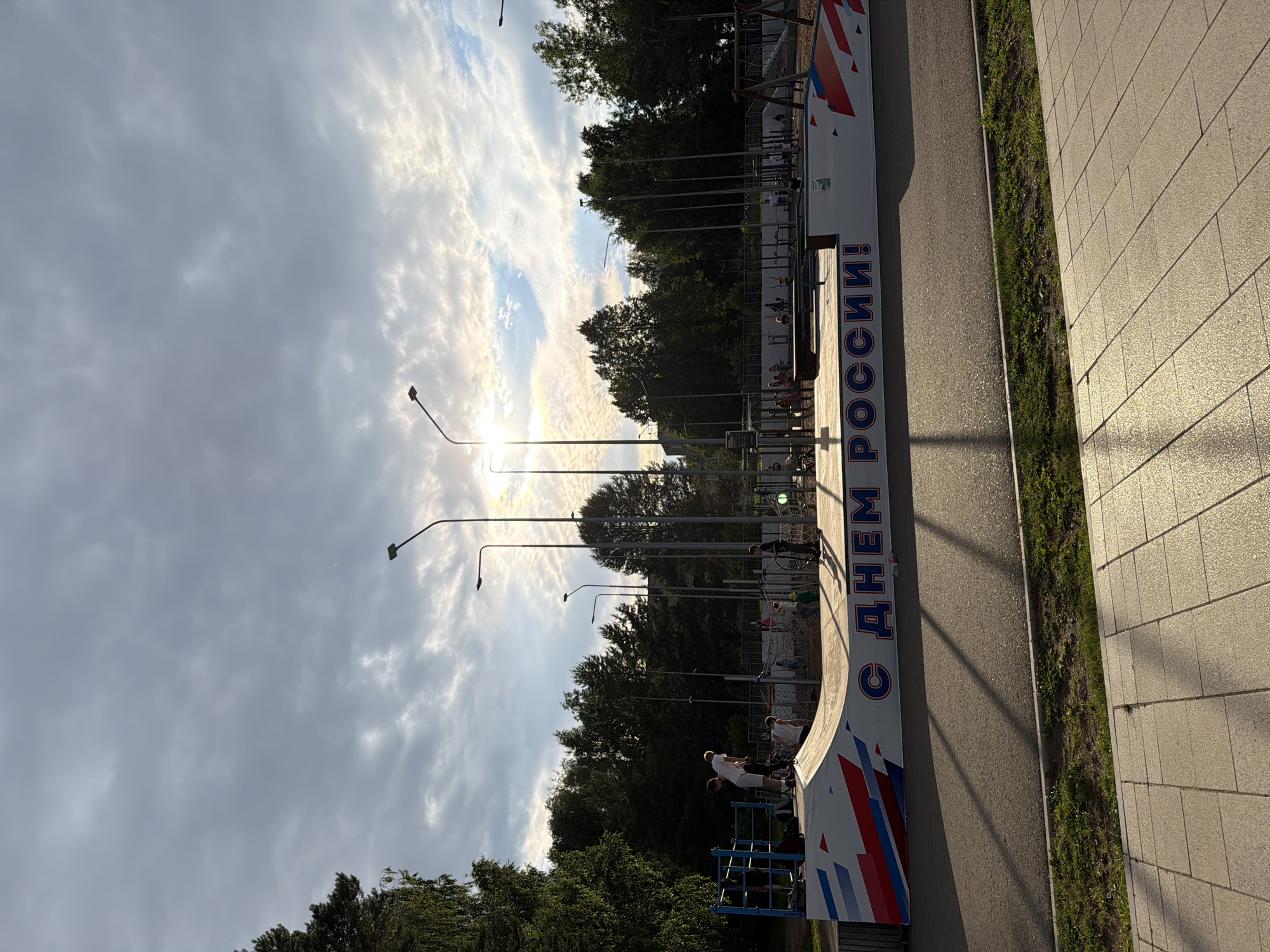 This media file captures a sunny day at an outdoor skatepark in Chaikovsky, Russia. The image features a concrete ramp with a white surface decorated with red, white, and blue geometric patterns. Large blue Cyrillic text adorns the side of the ramp, reading "С ДНЕМ РОССИИ!" which translates to "Happy Russia Day!".
Several people are present, engaged in activities typical of a skatepark. Some individuals are on skateboards, either preparing to ride the ramp or in the process of performing tricks. Others appear to be observing or waiting for their turn. The general atmosphere seems active and recreational.
The setting is an urban park with a paved walkway on the right side of the image. Tall streetlights are visible, interspersed with lush green trees that line the background. The sky is partly cloudy, with the sun breaking through, casting a warm, golden light that suggests it is late afternoon. The sunlight creates distinct shadows on the ramp and the surrounding area, contributing to a sense of depth and time. The overall impression is one of a community gathering in a public space to celebrate a national holiday, enjoying outdoor activities under pleasant weather conditions.