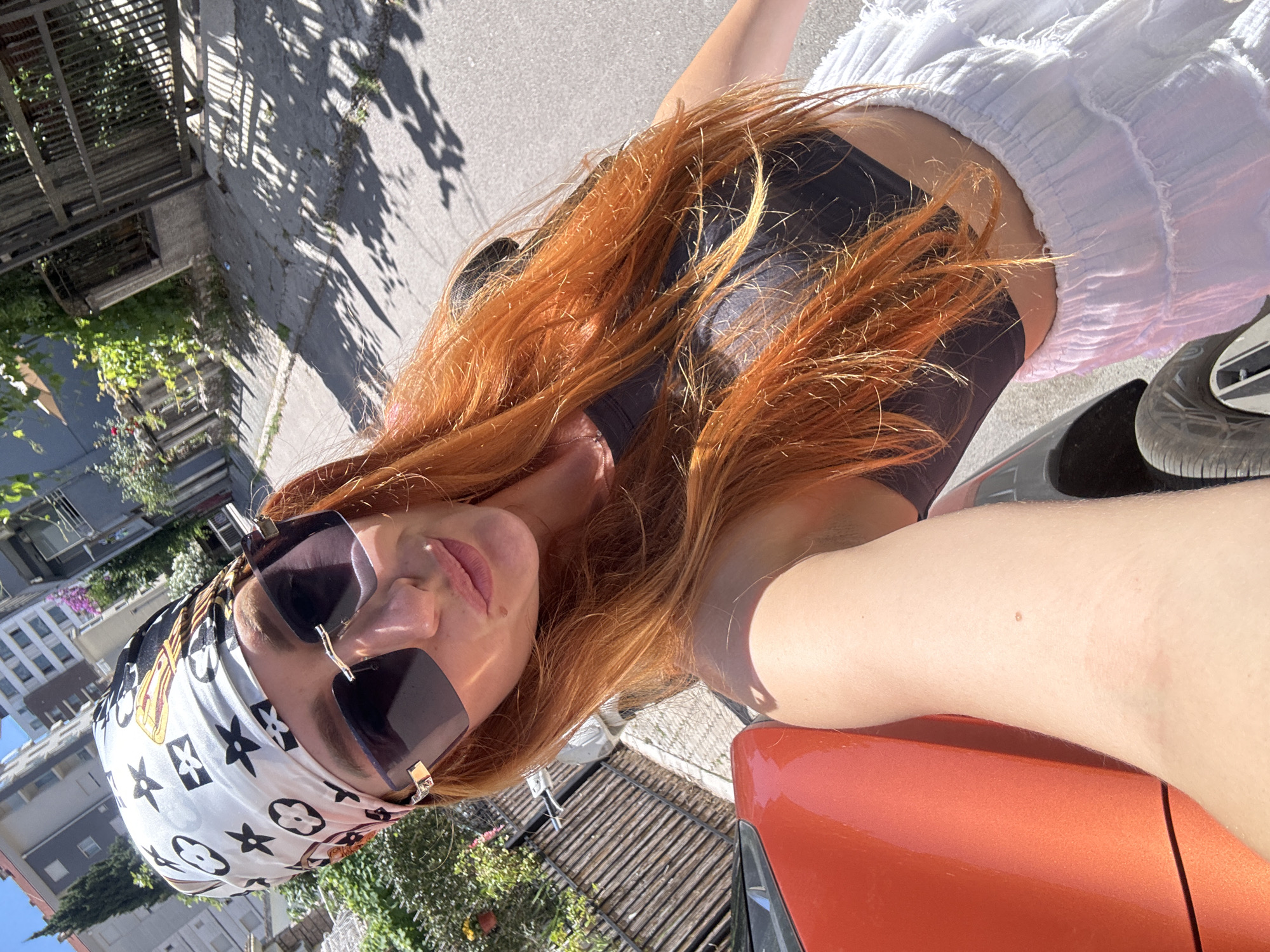 A woman with vibrant red hair, wearing a black crop top and a white ruffled skirt, poses for a selfie outdoors. She has a white scarf with a black and gold pattern tied around her head and is wearing oversized sunglasses. The background suggests an urban environment, with buildings and a street visible. The presence of an orange car in the foreground indicates she is either arriving or departing from this location. The sunny weather, indicated by the shadows on the pavement, suggests it is daytime. The scene appears to be in the city of Budva, Montenegro.