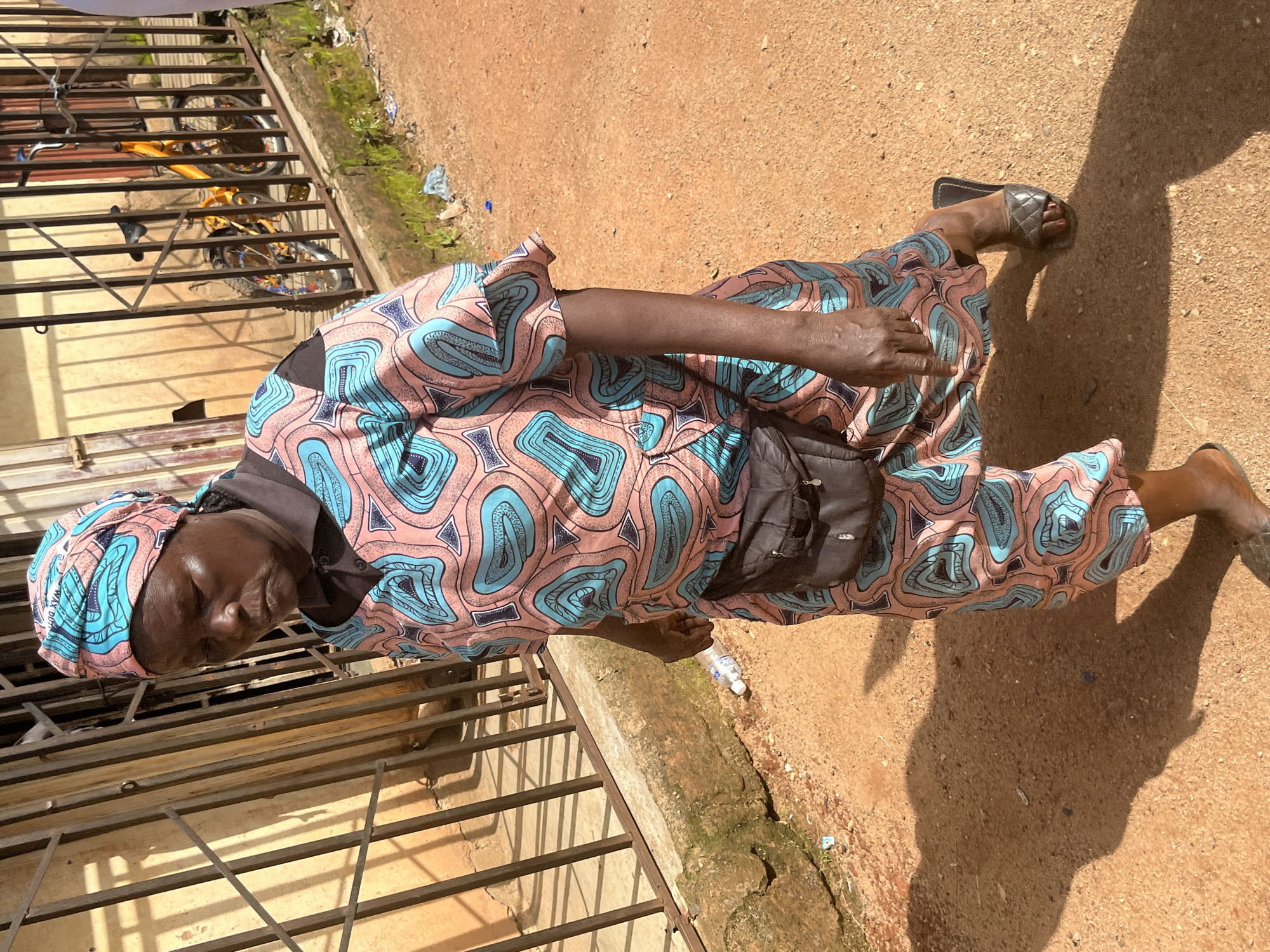 A woman is walking outside near a fence. She is wearing a colorful patterned dress and headscarf, a black fanny pack, and black sandals. The ground is dirt and there is a bicycle behind the fence. The scene appears to be in Jos, Nigeria.
