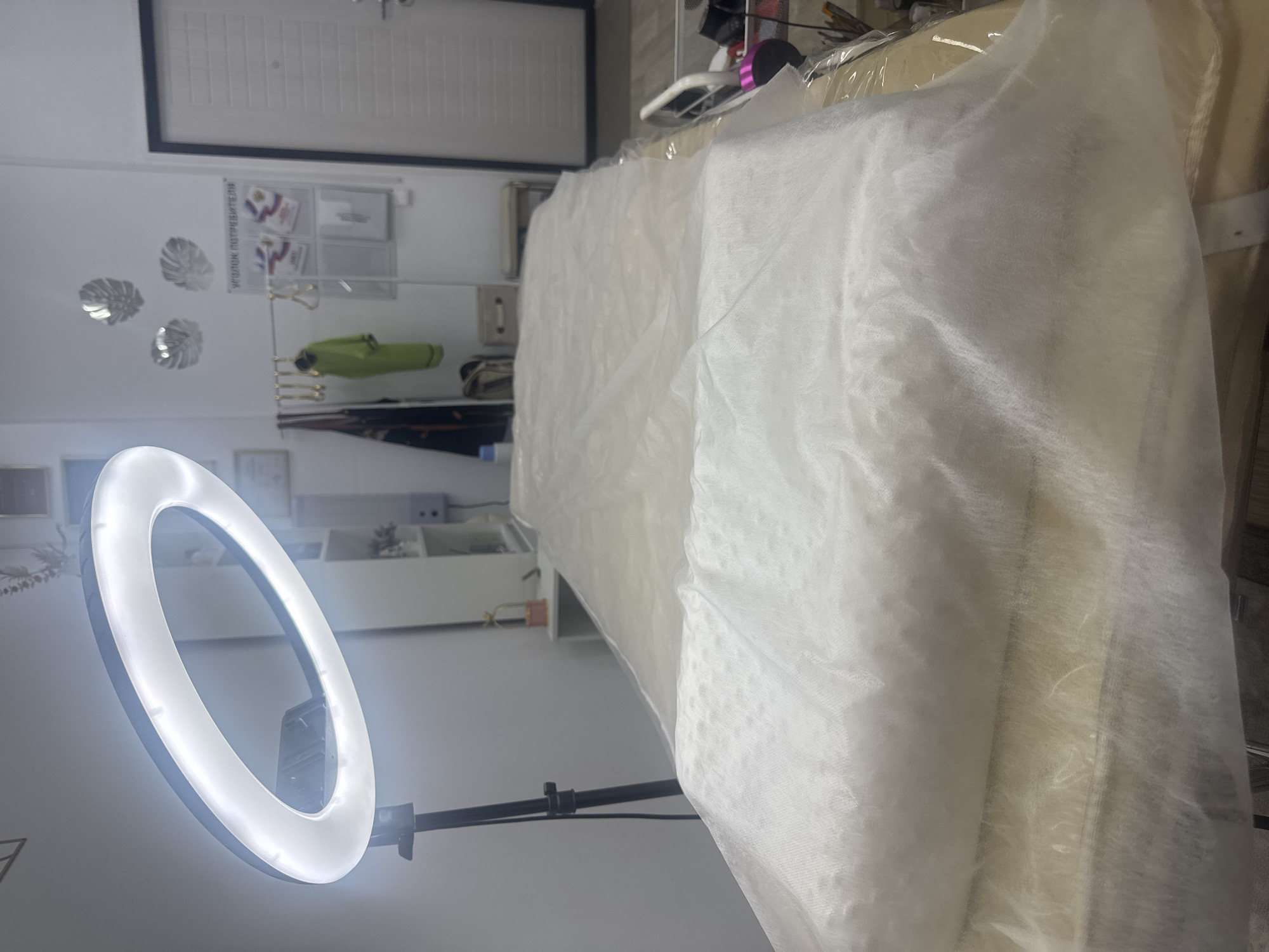 The image depicts a clean, well-lit room, likely a beauty salon or a similar establishment, located in Komsomolsk-on-Amur, Russia. A massage or treatment table, covered with a light-colored disposable sheet, is positioned in the foreground, dominating the scene. The sheet is wrinkled and appears to be made of a non-woven material, possibly for hygiene purposes. A ring light is visible in the upper left corner, mounted on a black tripod, indicating that the space is set up for professional use, possibly for photography or video recording during treatments.
In the background, a door with a white paneled design is prominent on the right side of the frame. To the left of the door, a wall displays decorative silver leaf-shaped wall art. A white notice board, labeled in Russian "Уголок потребителя" (Consumer's Corner), is mounted on the wall, with several documents displayed within clear plastic sleeves. To the left of this board, a metal rail holds several garments, including a bright green jacket. Shelving units are visible further back, with some items placed on them, including what appears to be a small decorative item with a gold cage-like structure. A light-colored storage box is also present.
The overall impression is one of an organized and hygienic space, prepared for client services. The lighting is bright, and the decor is minimalist. The presence of the treatment table and professional equipment suggests the room is used for services such as massage, cosmetology, or aesthetic procedures. The location in Komsomolsk-on-Amur, Russia, is noted based on the Russian language on the notice board.