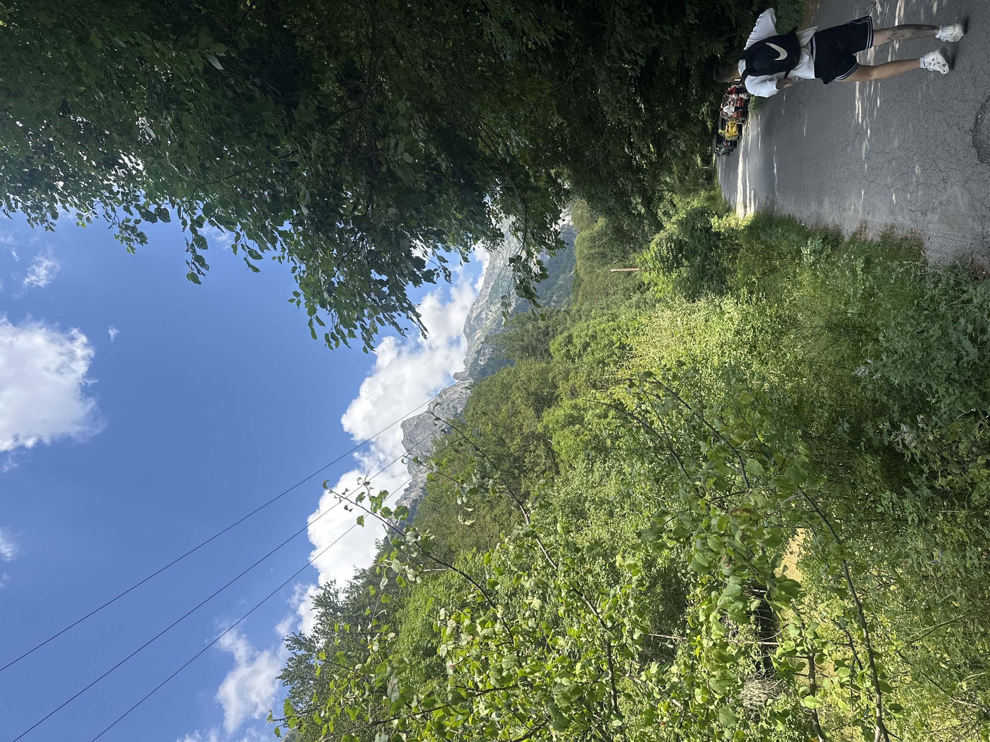 The image depicts a scenic road winding through a lush, green mountainous landscape. In the foreground, a young person wearing a white t-shirt, black shorts with white stripes, white sneakers, and a black backpack with a white Nike logo is running away from the camera on the paved road. To the left of the road, dense vegetation and trees cover a steep hillside, leading up to a majestic mountain range in the distance. The sky is a vibrant blue with scattered white clouds. Several cars are parked on the right side of the road, further down from the running figure. The overall impression is one of outdoor recreation in a beautiful natural setting, possibly near Rila Monastery, Bulgaria, given the mountainous terrain and the presence of a road and parked vehicles.