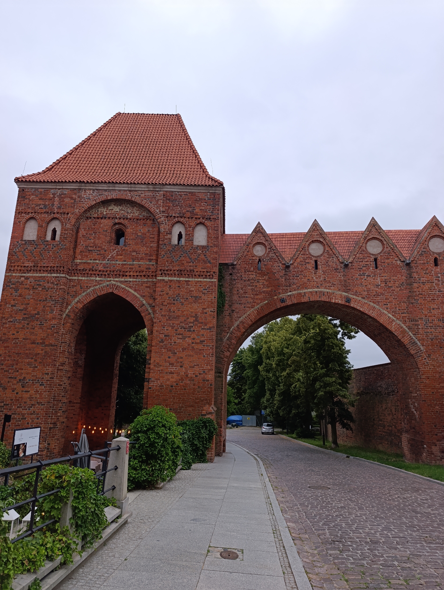 The image shows a view of the Toruń city walls in Poland, specifically the area around the bridgehead gate, which is a historical monument. The gate is constructed of red brick and features a large arched entryway. Adjacent to the gate is a cobbled road, and to the left, a sidewalk with a railing and ivy-covered planters. The sky is overcast. The gate leads to a street lined with trees.
