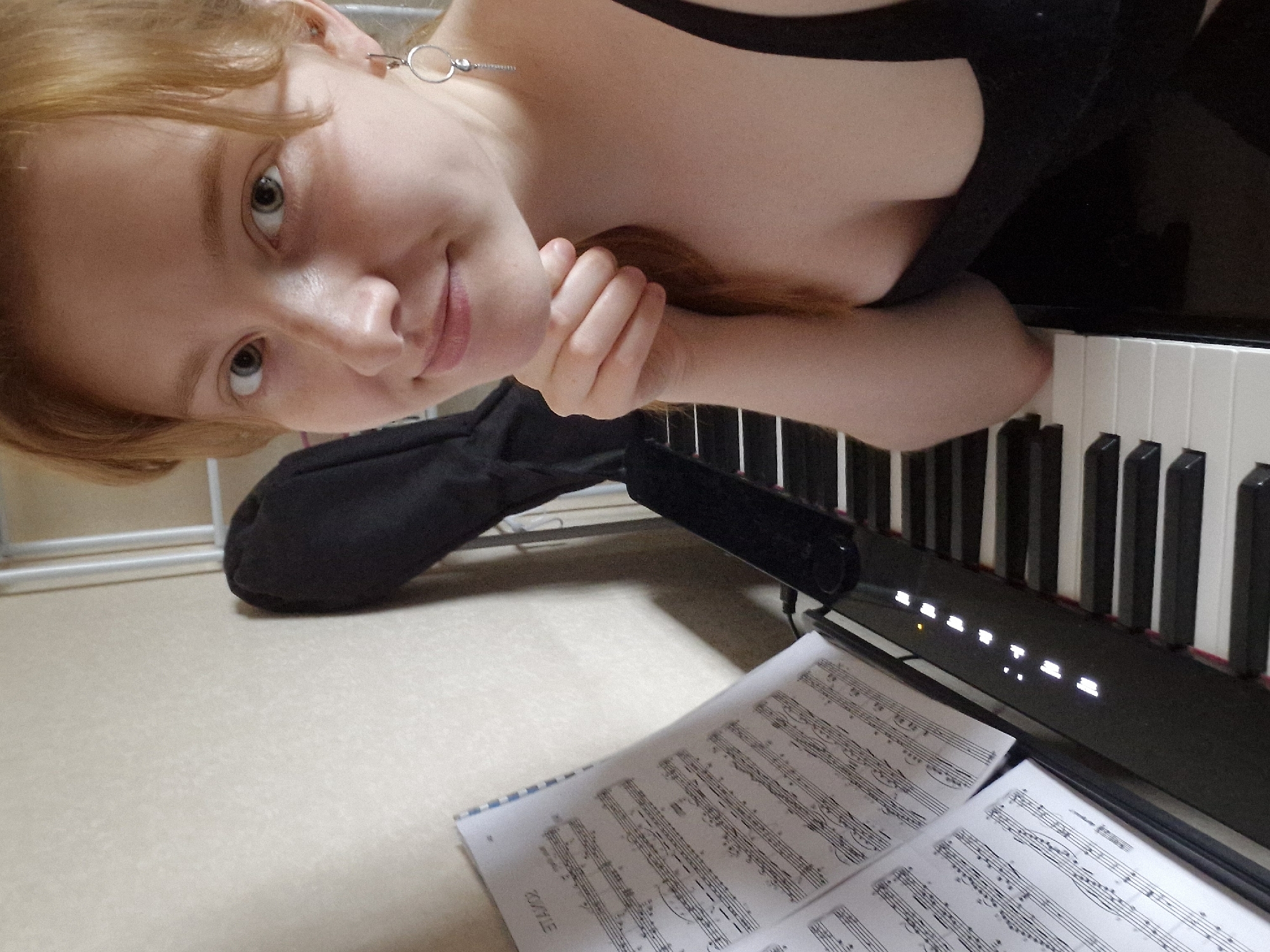 A fair-skinned young woman with reddish-blonde hair is positioned in front of a digital piano. She is wearing a black tank top and has an earring. Her left hand is resting on top of her right, near her cheek. Sheet music sits in front of the piano keys. A black bag or case is visible in the background. The background appears to be a room with light-colored surfaces. The location is not discernible from the image and cannot be determined as Yekaterinburg, Russia.