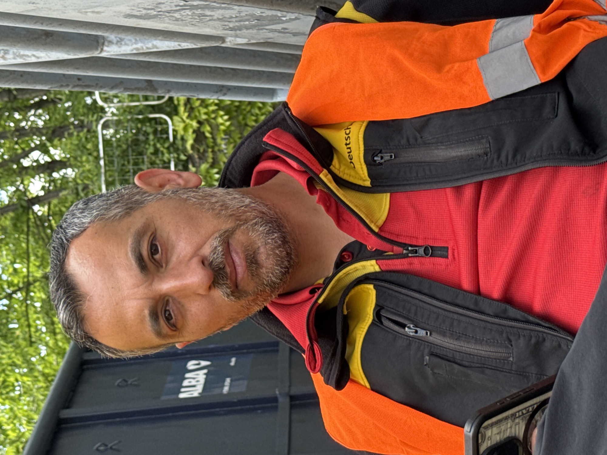 A man is seated outdoors, potentially in the city of Bietigheim-Bissingen, Germany, based on the "Deutsch" lettering on his jacket. The man has graying hair and a beard. He is wearing a red shirt under a black jacket with orange and gray reflective panels. The jacket has yellow accents near the collar and zippered pockets. Part of a container with the word "ALBA" is visible in the background, along with trees and an overhead metal structure. The man is holding a cell phone in his hand.