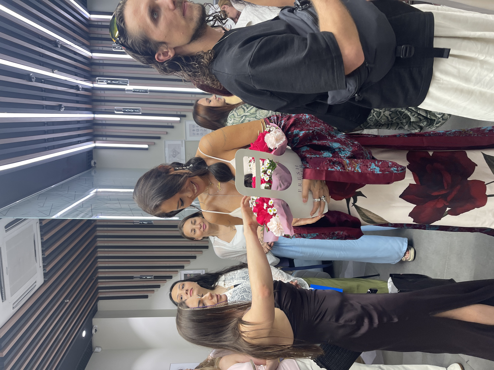 This image captures an indoor social gathering or event in Moscow, Russia, featuring several individuals in a modern commercial space.
In the center, a woman with long dark hair, wearing a white spaghetti-strap top, a red floral patterned skirt, and a patterned dark red and teal jacket, smiles as she receives a bouquet of red roses and white/pink carnations. The flowers are held in a white container labeled "FRESH FLOWER." A woman to her left, with long dark hair and wearing a dark brown strapless dress, extends her right arm to present the bouquet. She has a decorative element near her left eye. To the far right, a man with long, curly dark hair, wearing a black t-shirt and a black fanny pack, looks towards the right of the frame. He has sunglasses on his head.
In the background, other individuals are visible: a woman behind the flower giver, a woman in a white shirt and light blue wide-leg pants smiling faintly, and several partially obscured figures.
The setting features white walls adorned with framed illustrations of diamond shapes, suggesting a jewelry atelier or similar retail space. The ceiling is composed of alternating dark and light-toned slatted panels, interspersed with recessed strip lighting and several hanging cylindrical pendant lights. A square HVAC unit is visible on the ceiling in the upper left, along with a small security camera. A glass partition with etched details is present on the right side of the frame. The overall atmosphere appears bright and social.